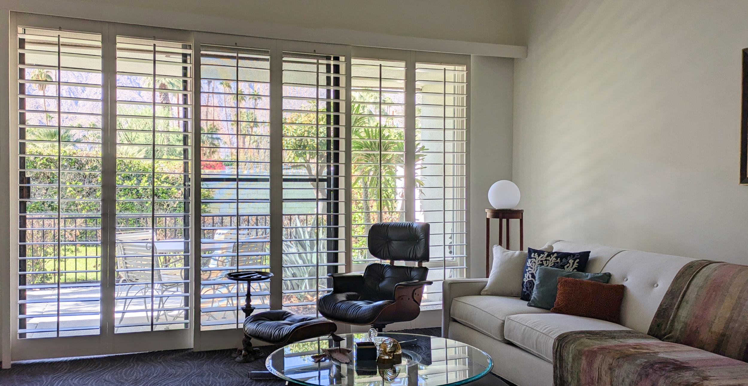 Living room with large windows with white blinds, a black lounge chair with a matching ottoman, a cream sofa with decorative pillows, a glass-top coffee table, and an outdoor patio visible through the windows.