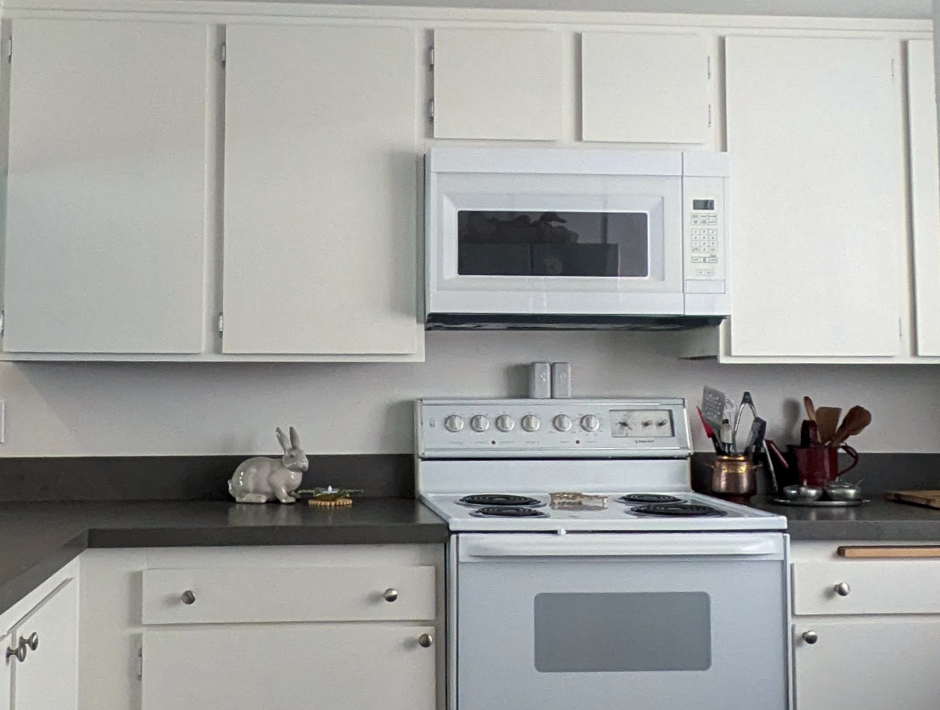 White kitchen with white cabinets, black countertop, microwave above stove, Rabbit figurine, cooking utensils, and small containers on the counter.