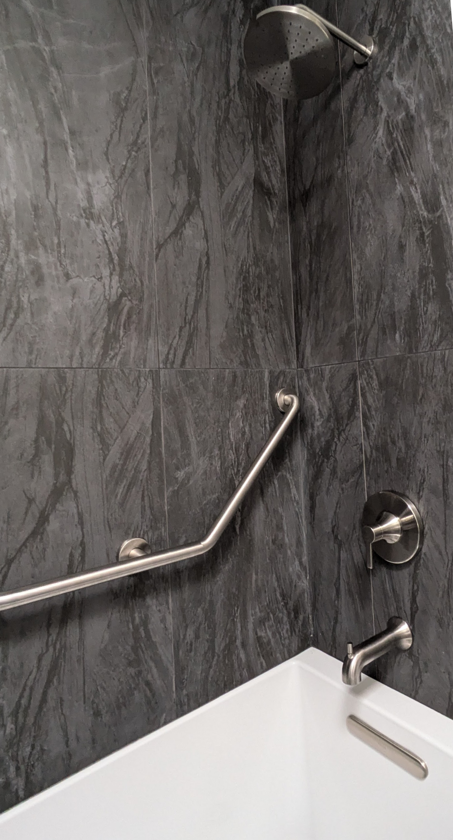 Shower with gray marble tiles, metal grab bar, overhead showerhead, and silver faucet.