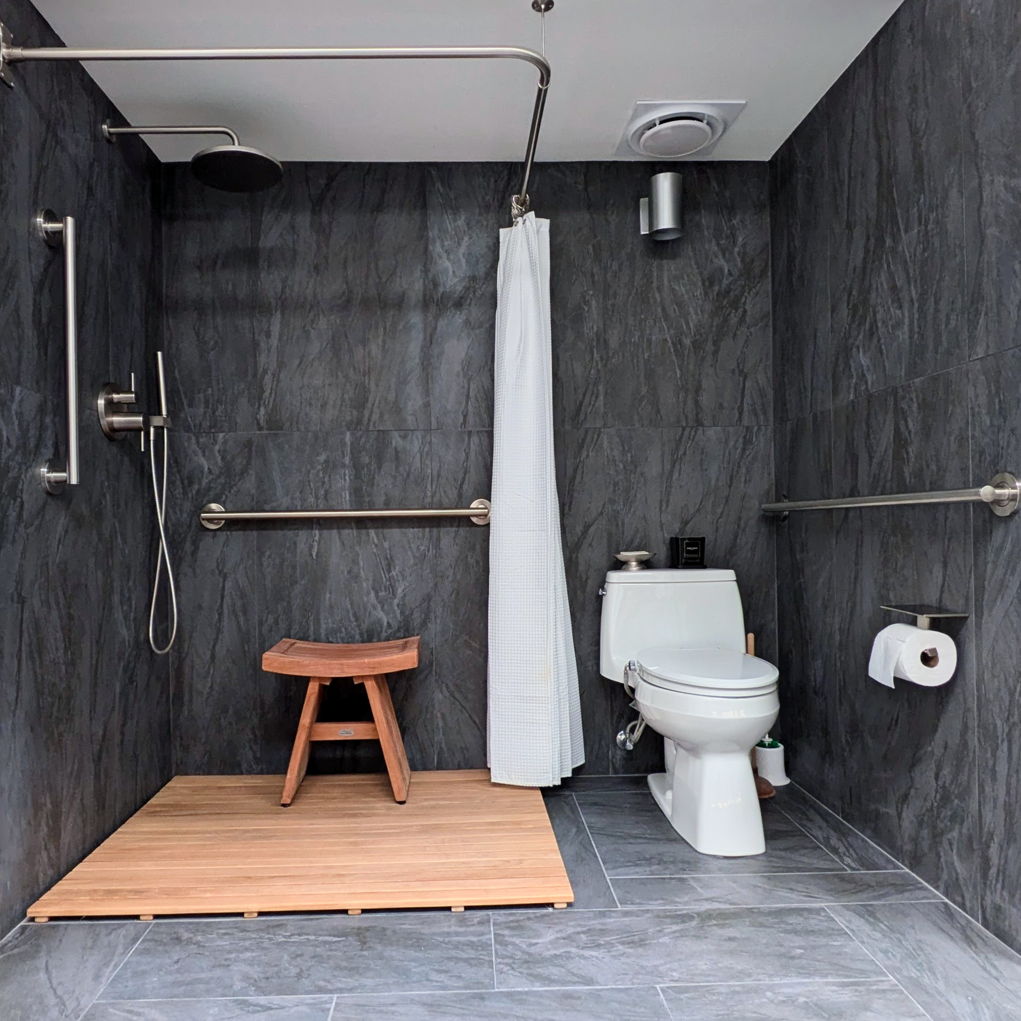 Accessible bathroom with dark gray tiled walls and floor, a walk-in shower with a wooden platform, a showerhead, a hand-held shower, two grab bars, a wooden stool, a white toilet with a black box on top, a toilet paper holder, a small plant, and a white shower curtain.