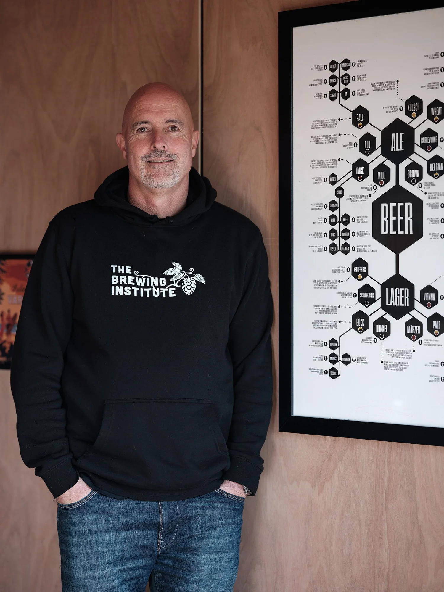 A man in a hoodie standing beside a framed brewery poster