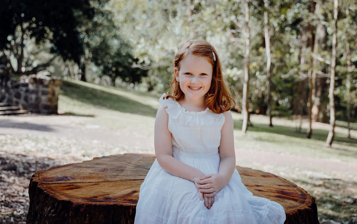 A young girl in a white dress sits on a tree stump in the park