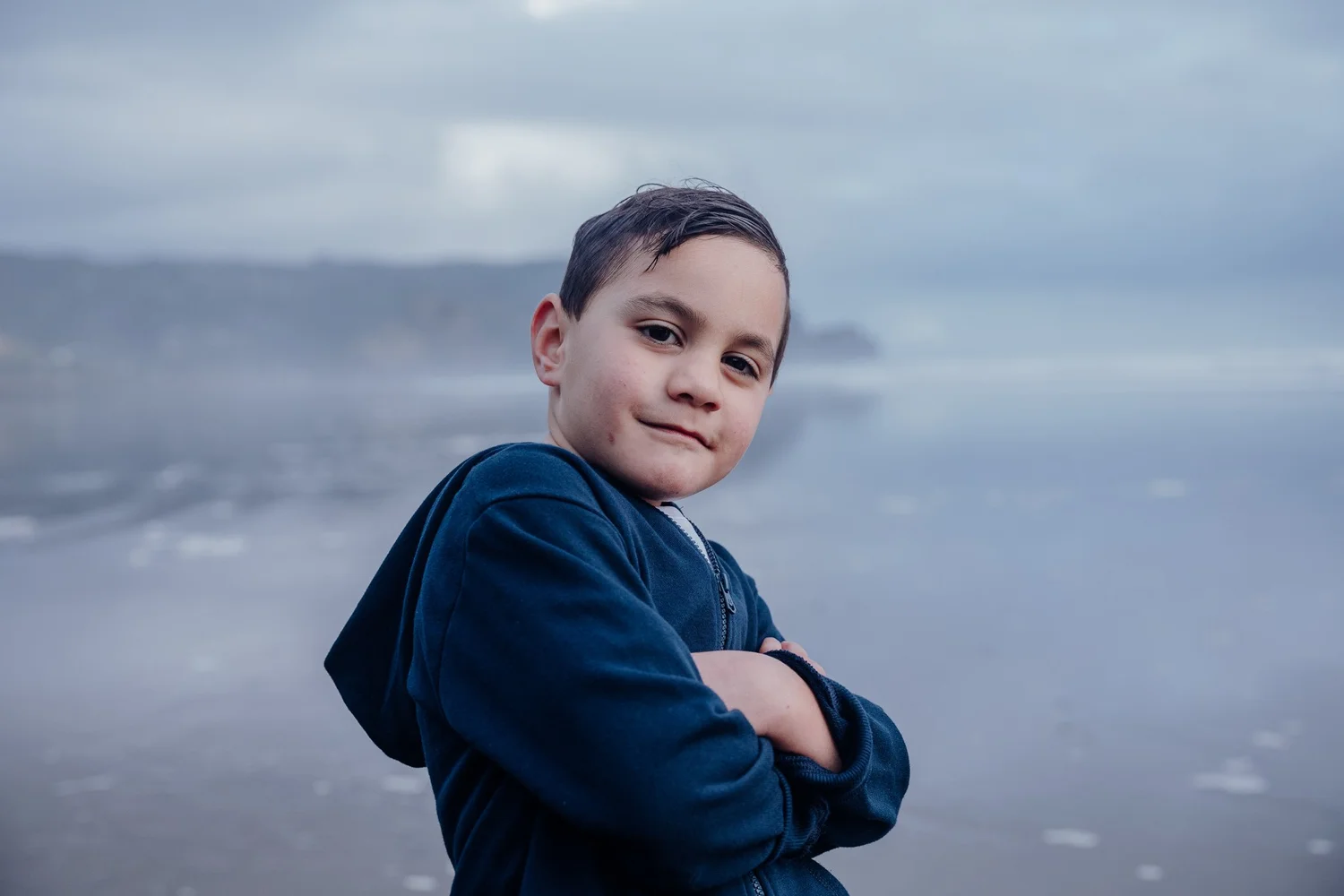 A boy in a hoodie poses with folded arms by the sea