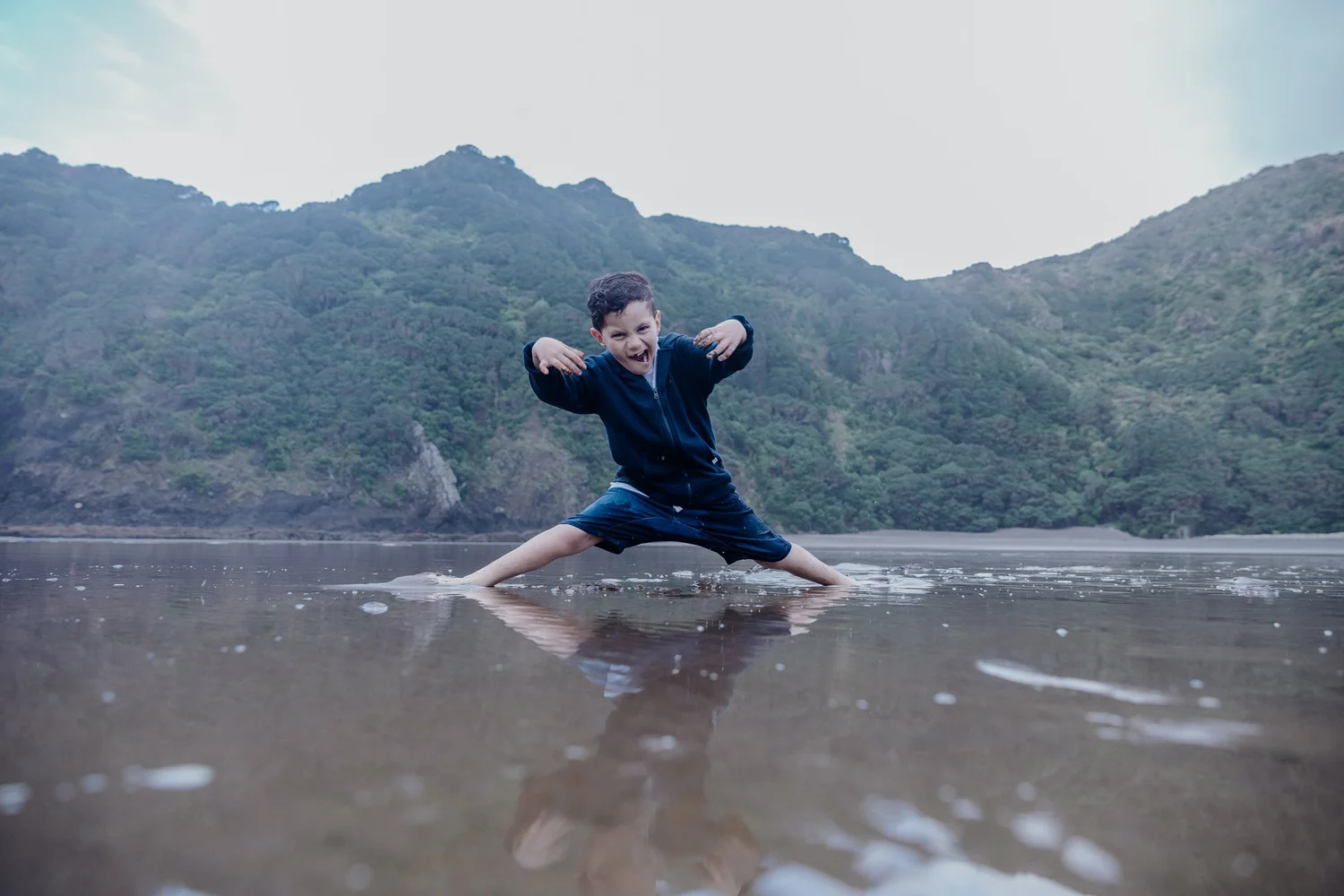 A boy strikes a playful pose on the sand with hills behind