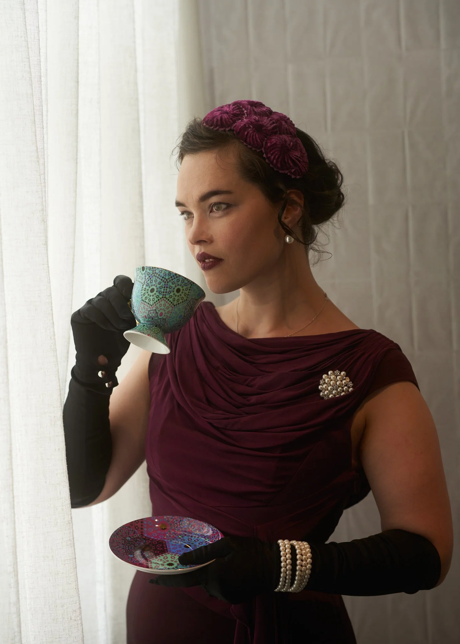 Anita sips from a teacup in burgundy and black gloves