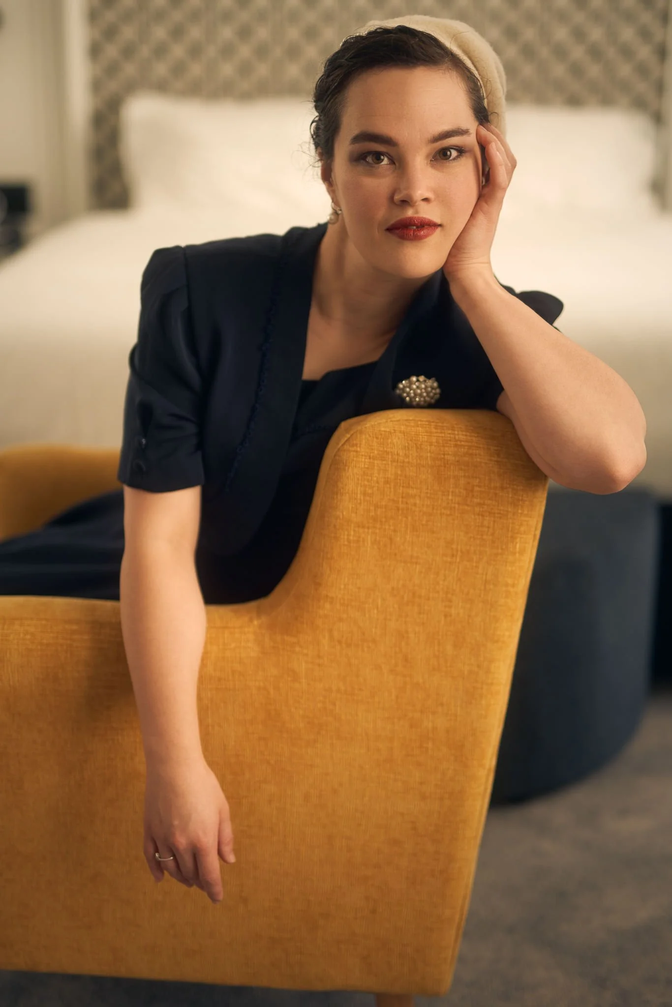 Anita in a navy dress resting her head on a mustard armchair