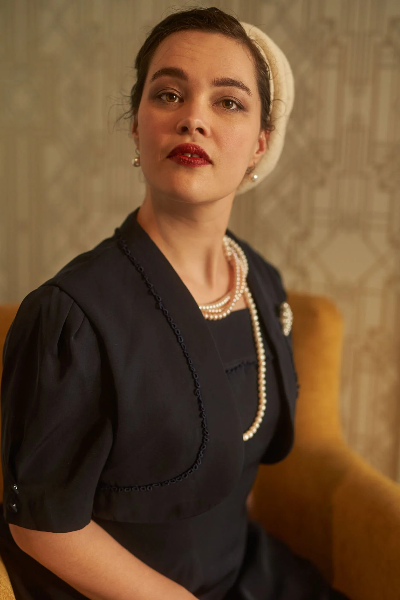 Anita in a cream beret and pearls on a mustard armchair