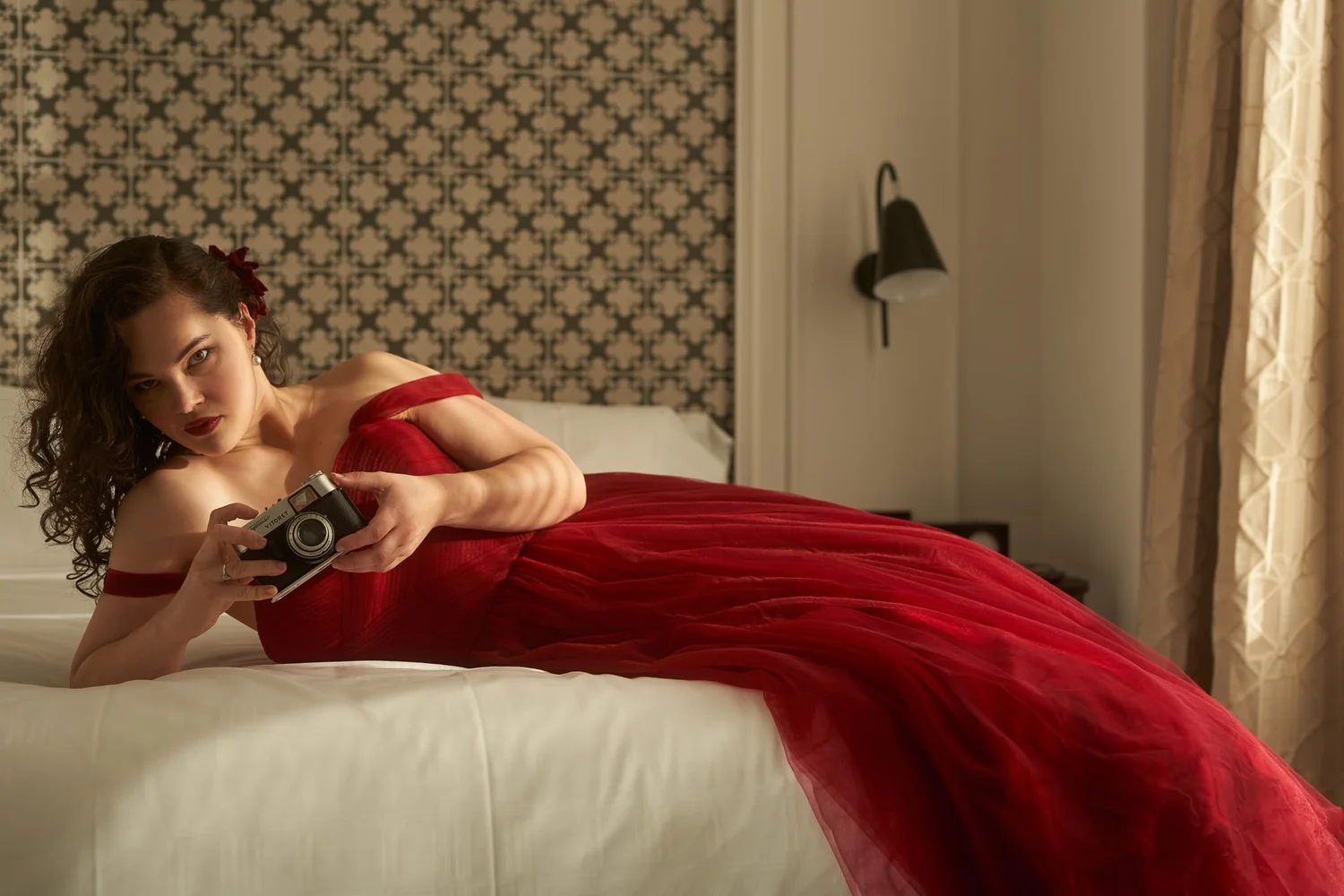 Anita in a red tulle gown lying on a hotel bed at The Airedale with a vintage camera