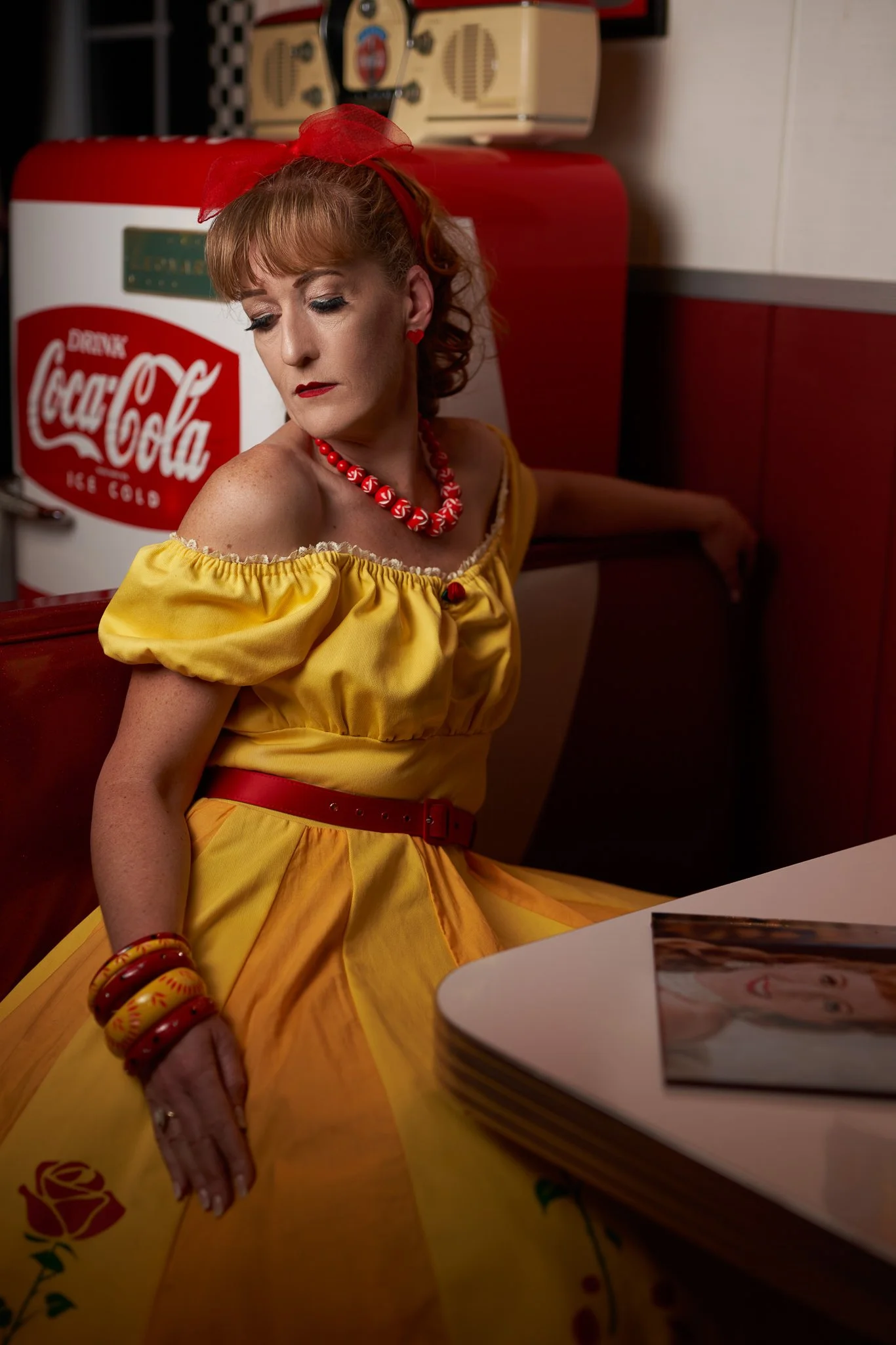 Sandy in a yellow 1950s dress beside a vintage Coca-Cola cooler in her American diner