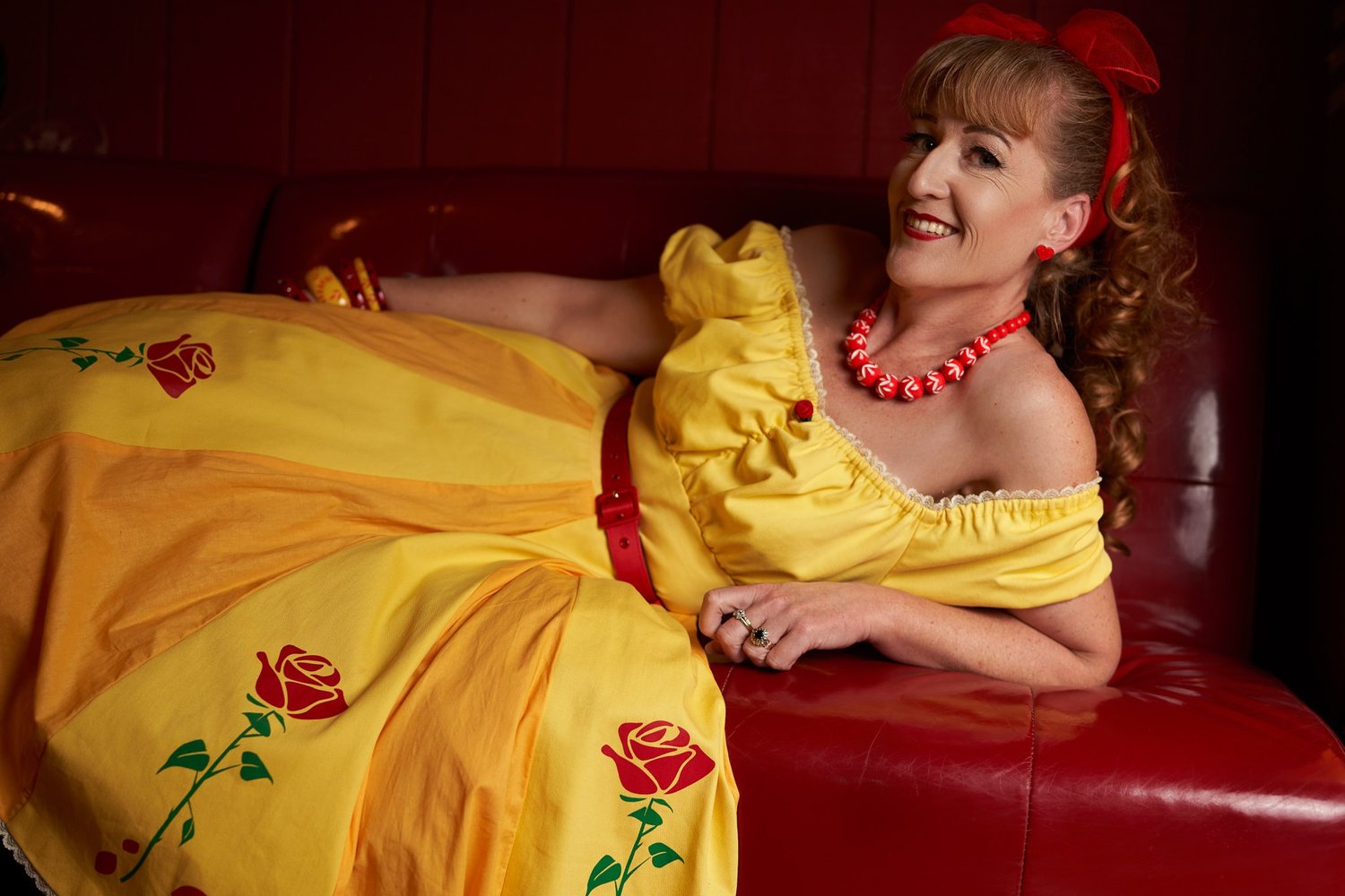Sandy reclines across a red booth in a yellow rose-print dress