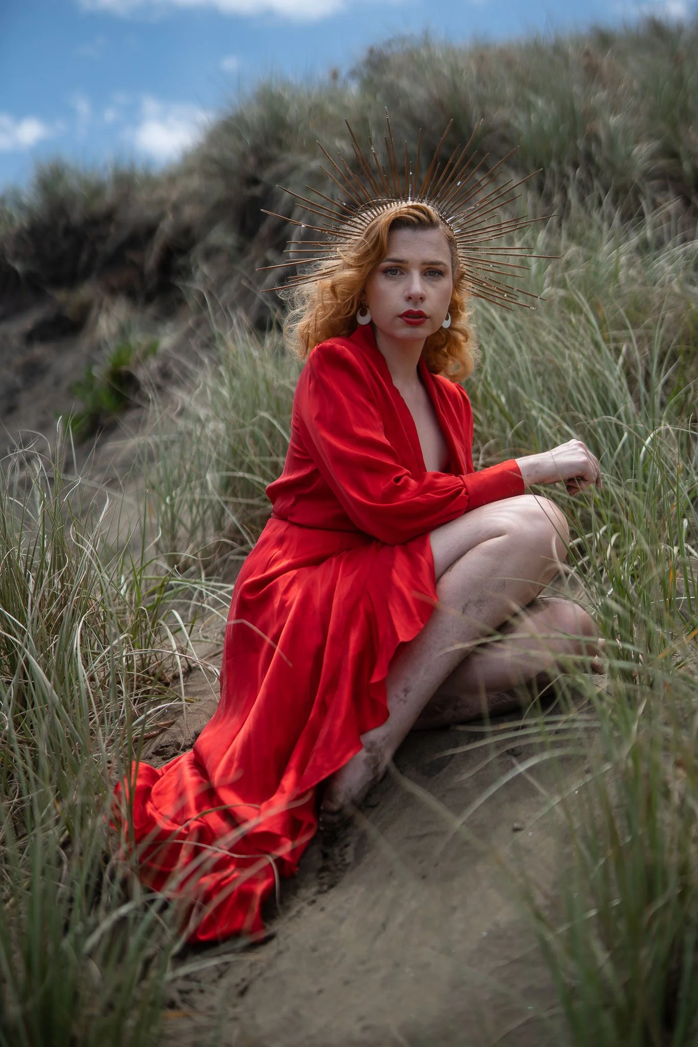 A model in a flowing red dress crouches on a sandy dune path