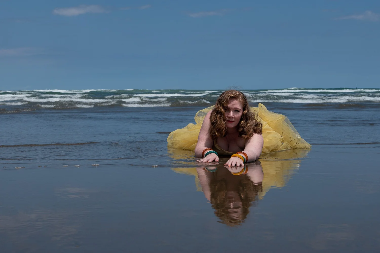A model in a yellow tulle dress lies on the wet sand at Muriwai