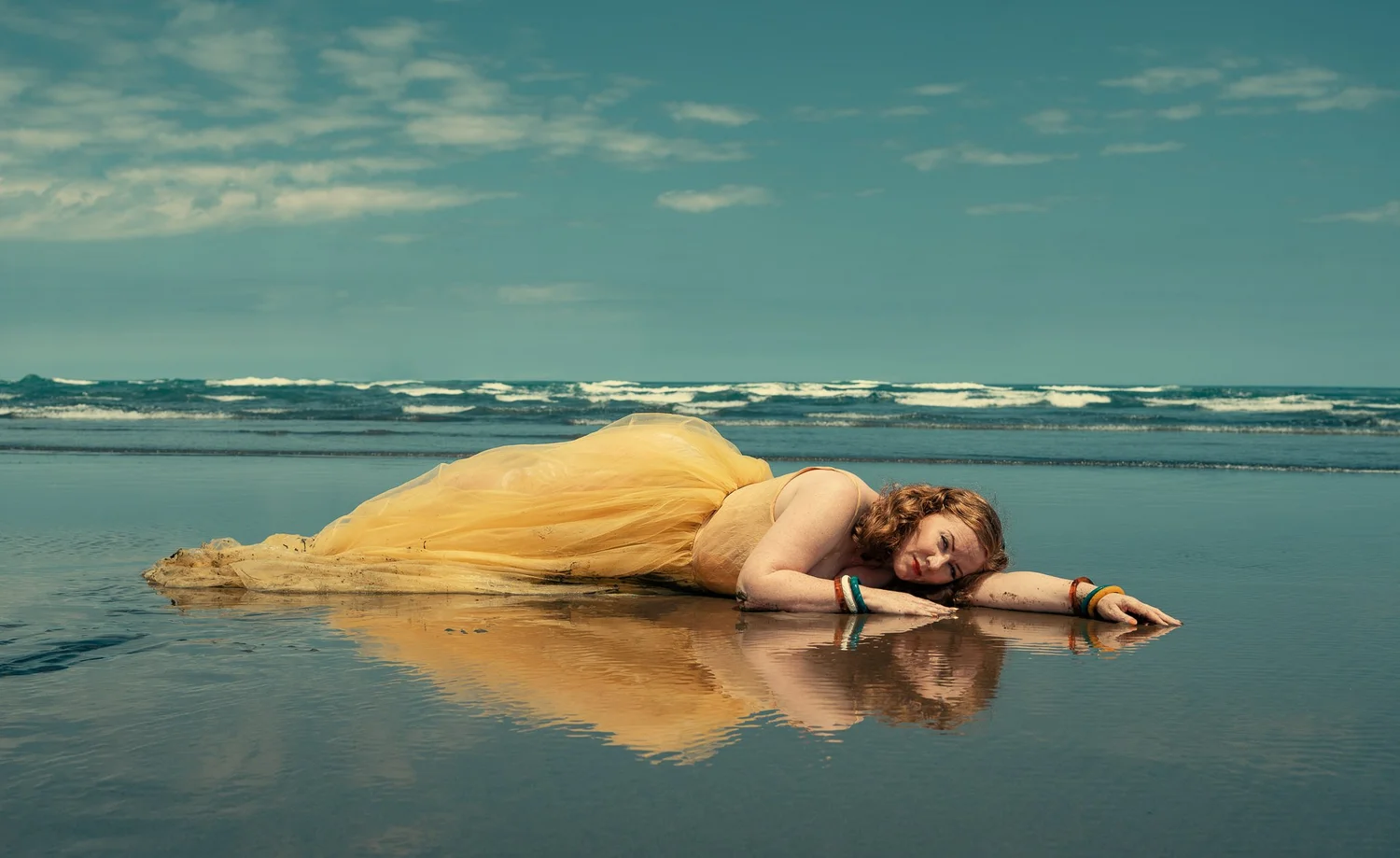 A model in yellow lies on the reflective sand by the surf