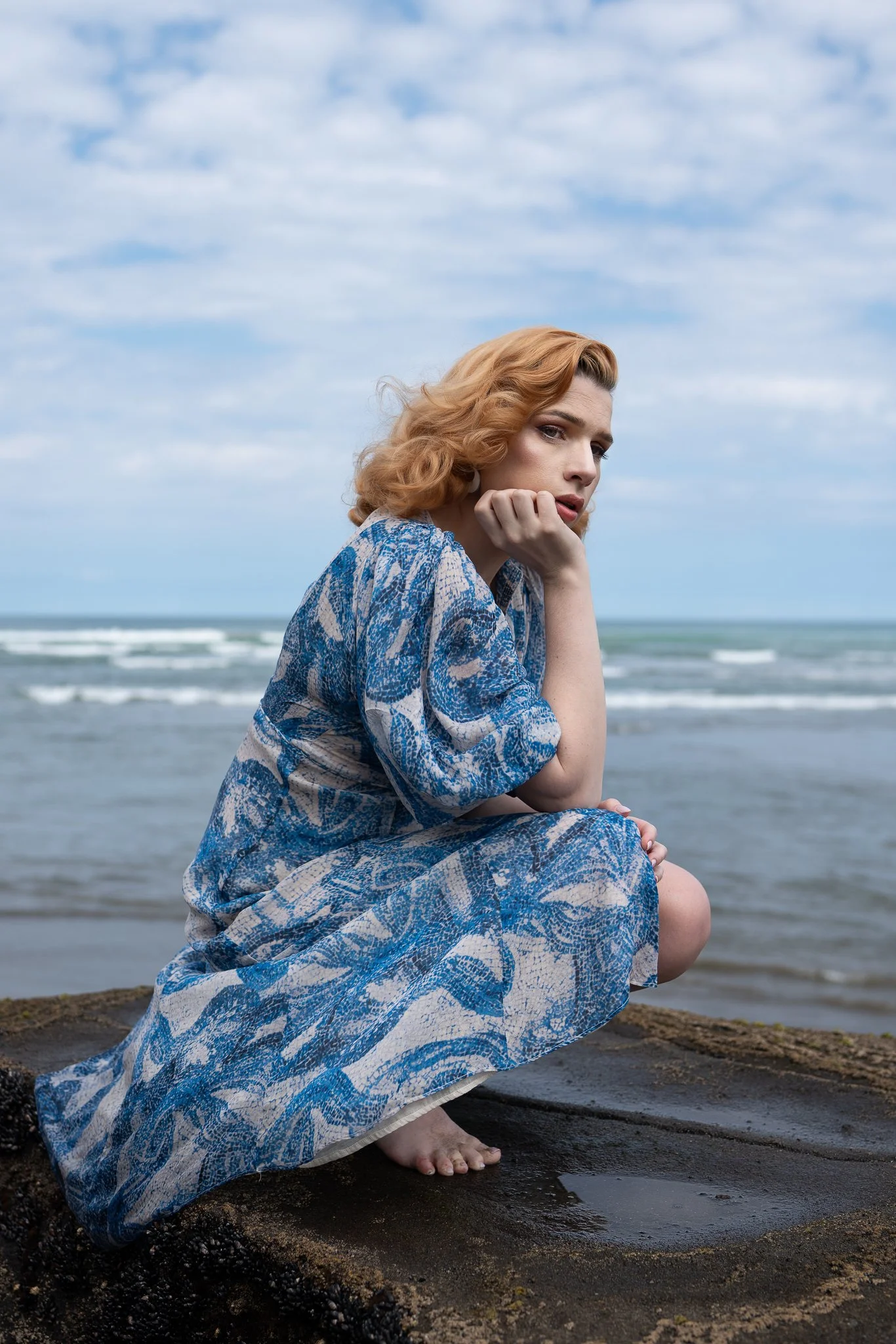 A model in a blue dress crouches on rocks by the sea