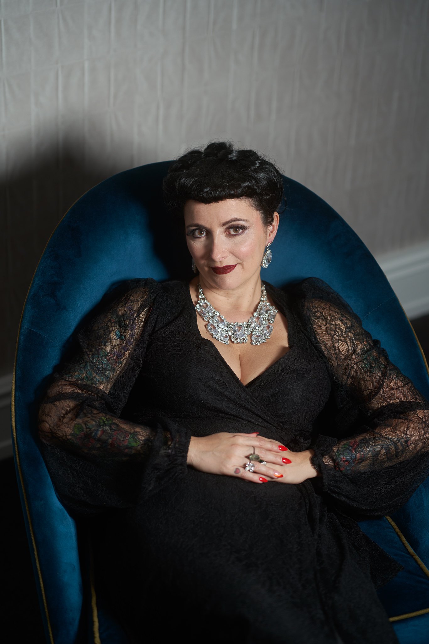 Viv in a teal velvet armchair wearing a diamanté necklace