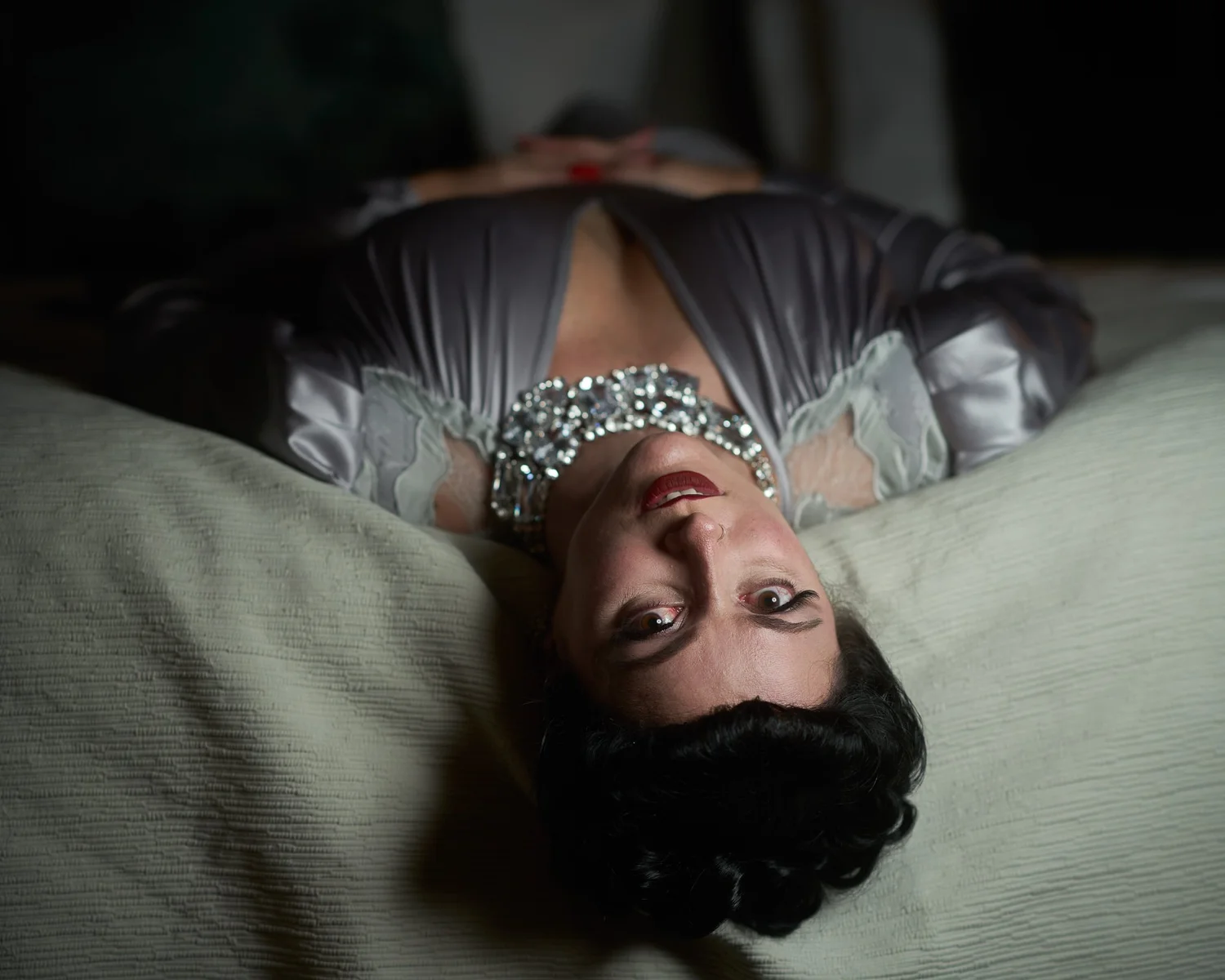 Viv lies upside down on a bed in a silver satin gown