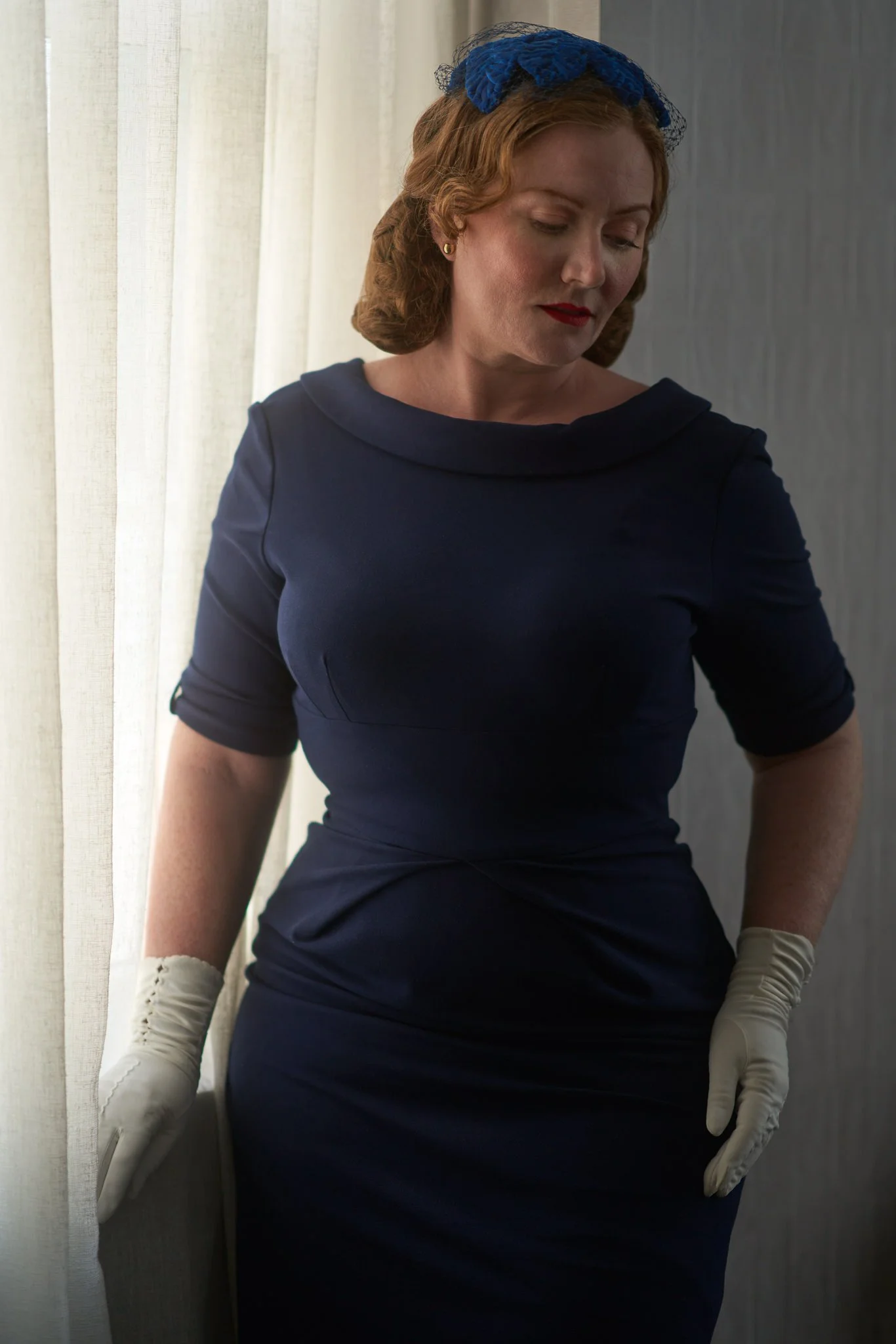 Laura in a navy dress and white gloves by a window