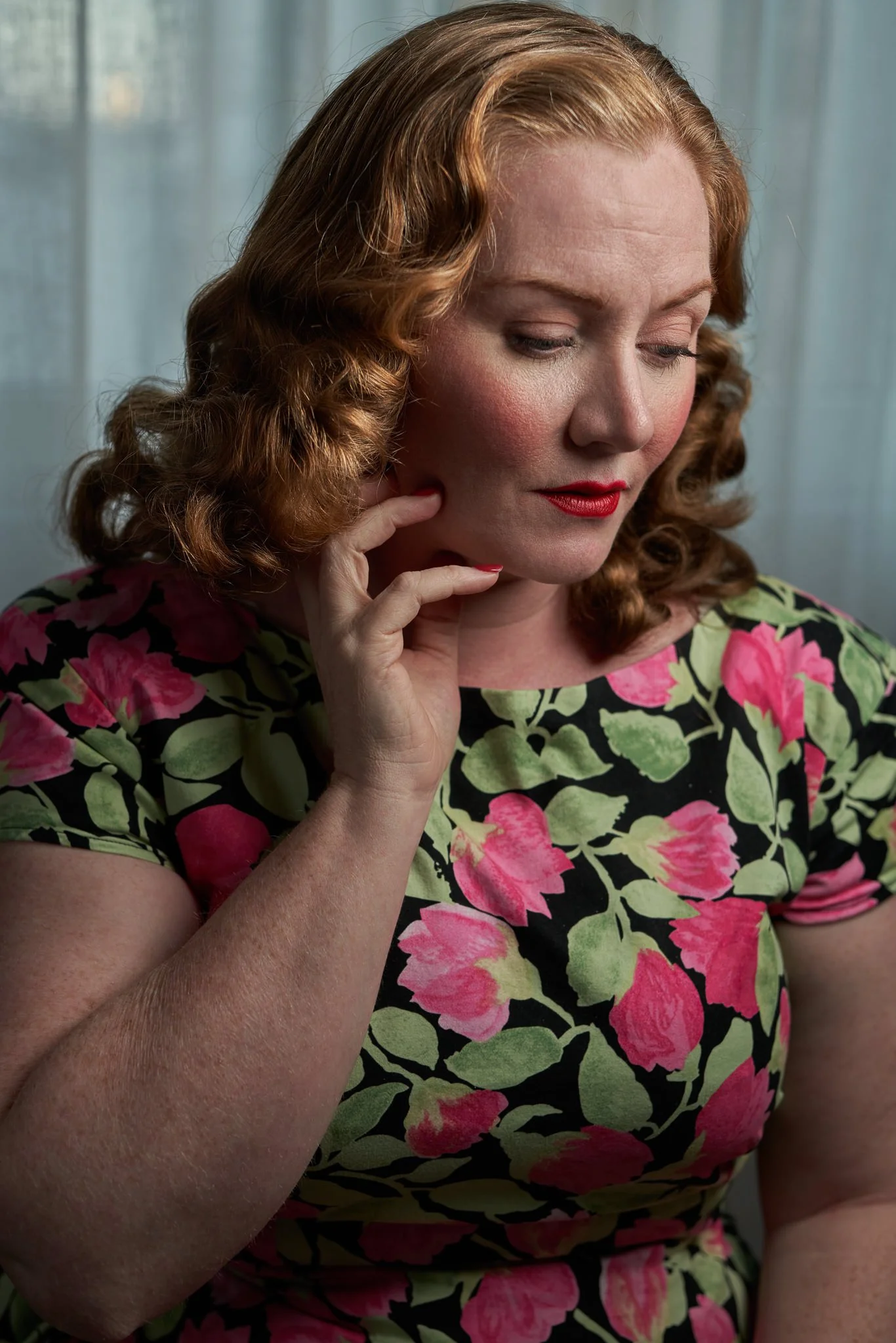 Laura in a floral dress looking down with a hand at her cheek