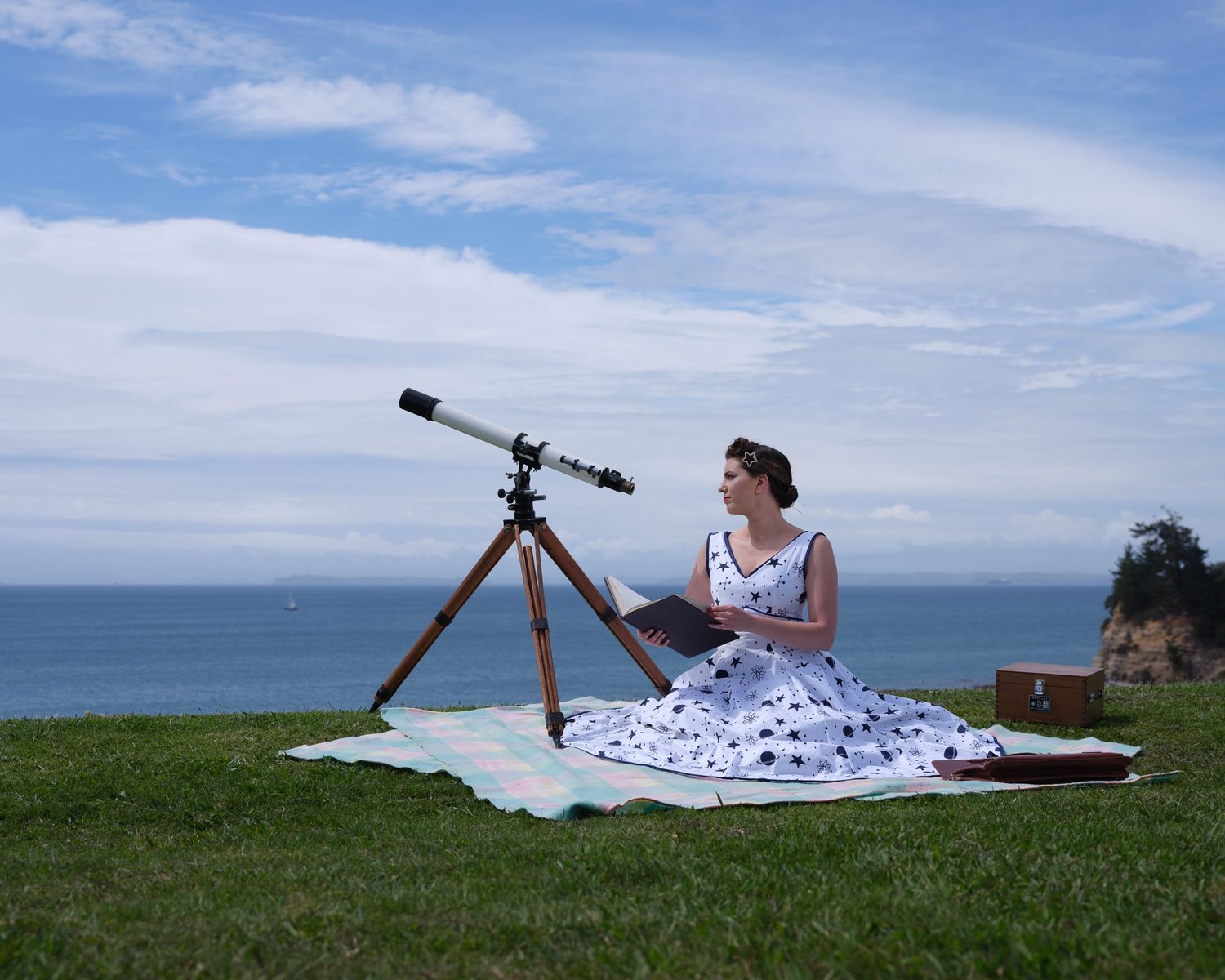 Jay in a polka dot dress on a picnic blanket beside a telescope by the sea