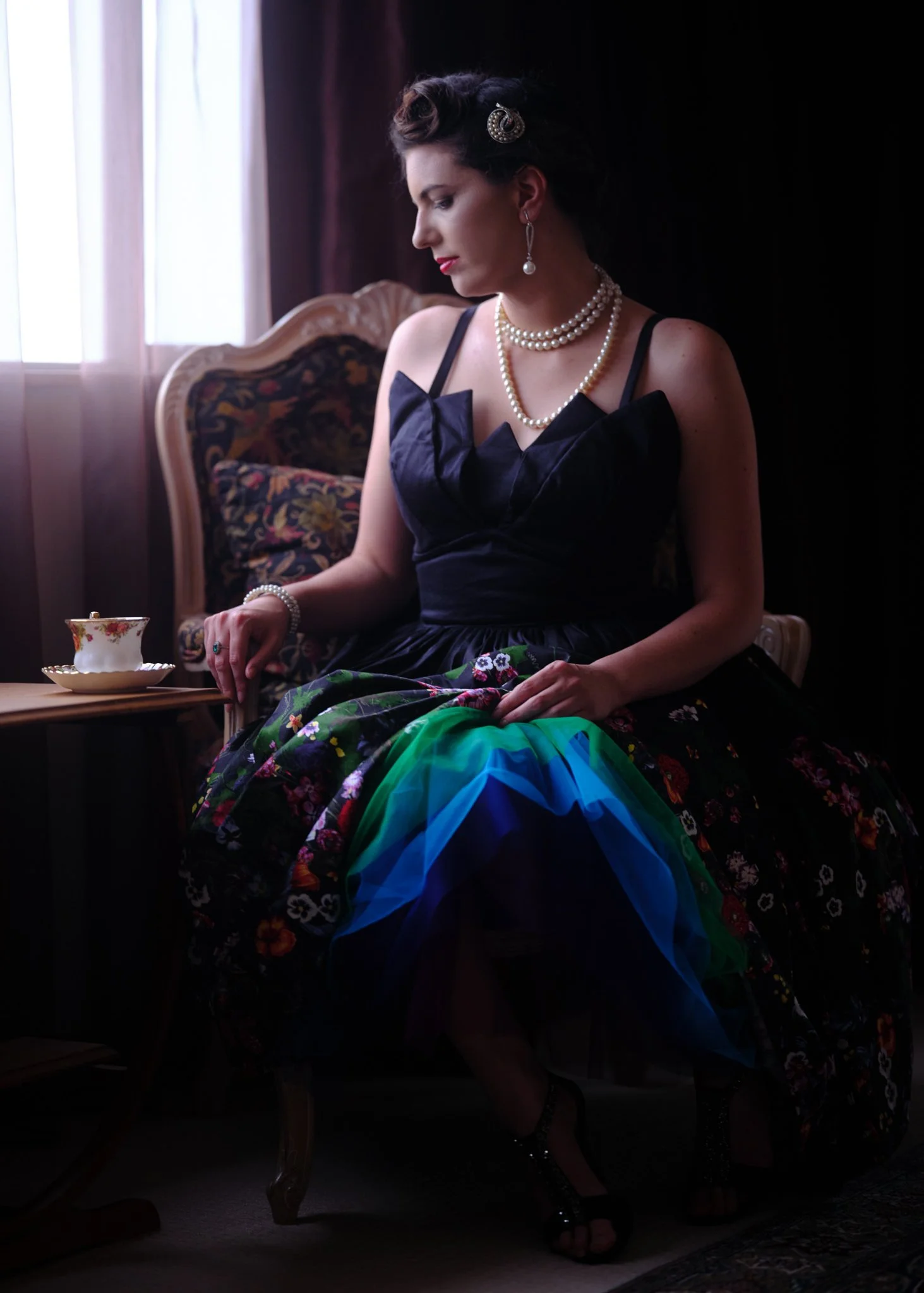 Jay seated by the window in a dress with a teal and blue skirt