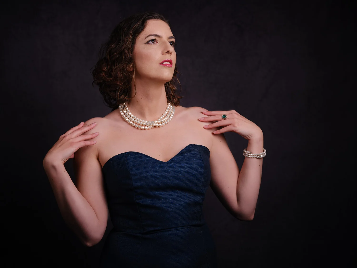 Jay in a strapless navy dress and pearls against a dark backdrop