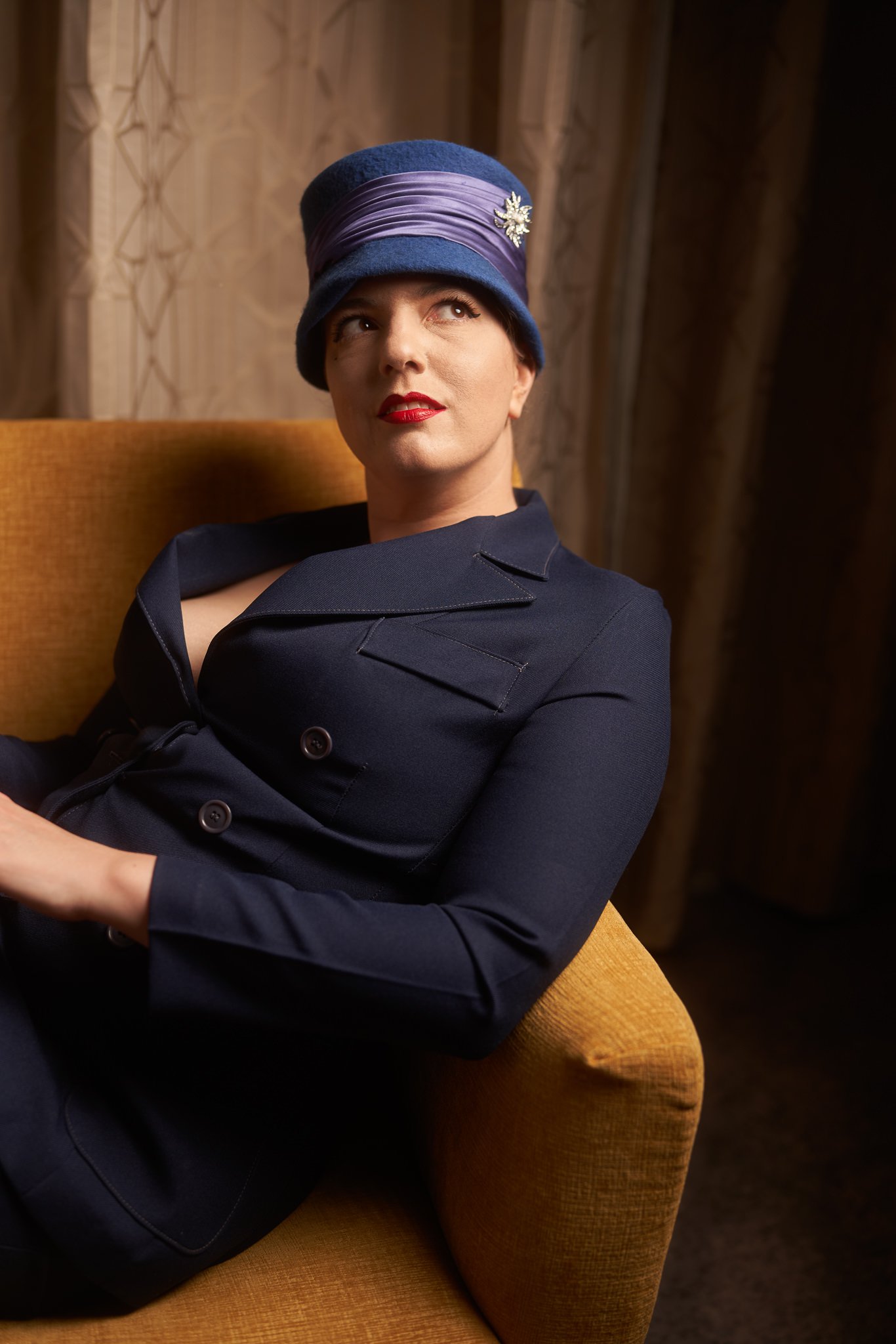 Jay in a blue cloche hat and navy jacket on a mustard armchair