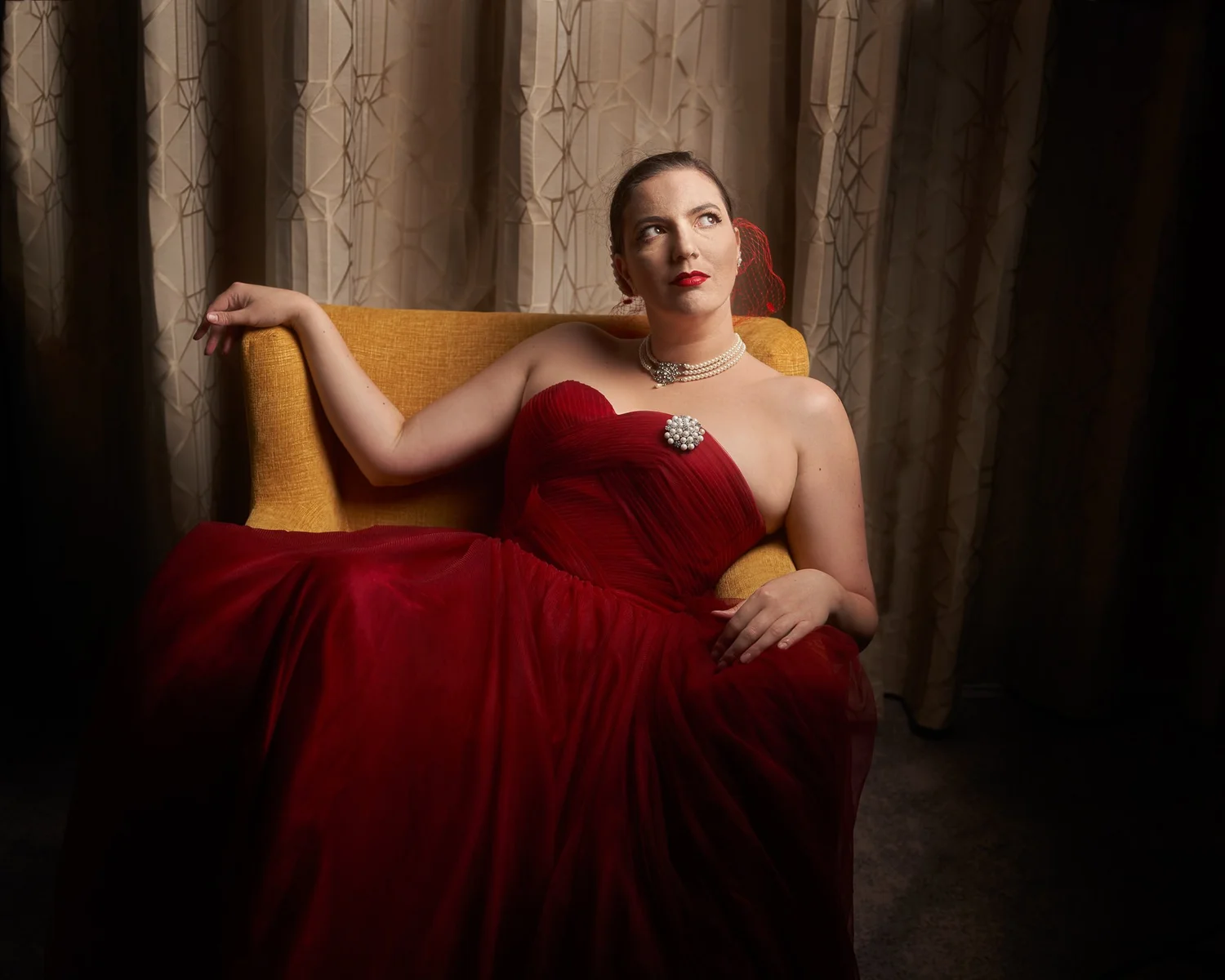 Jay in a red gown gazes upwards from a mustard armchair