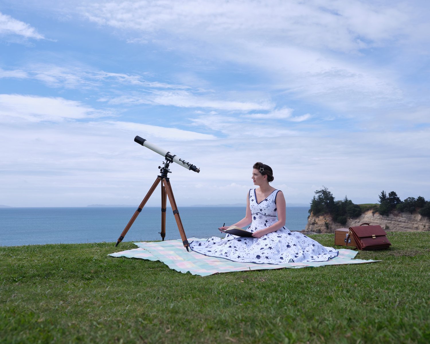 Jay reads on a picnic blanket beside a telescope on a clifftop