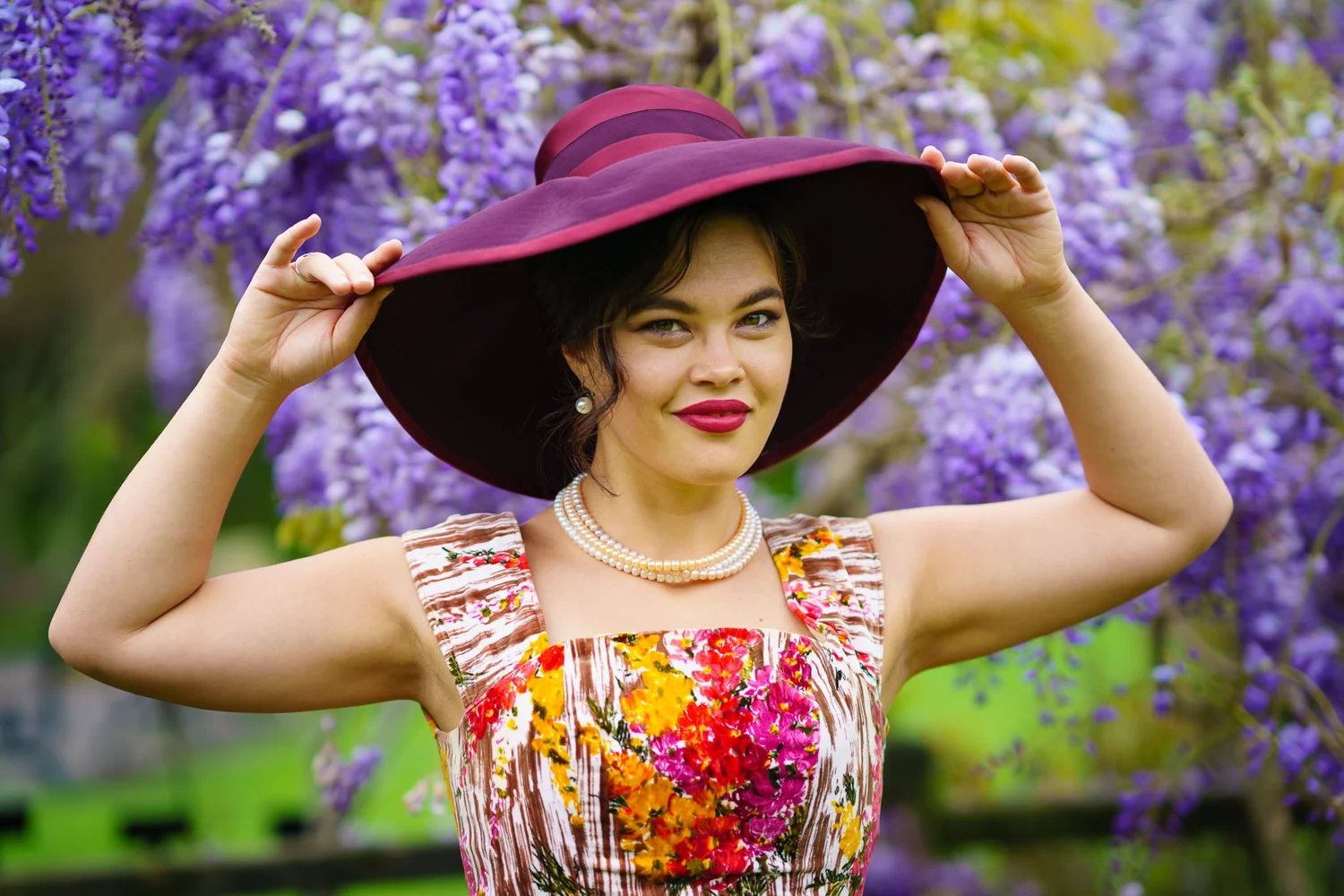 Anita in a wide-brimmed hat and floral dress under purple wisteria