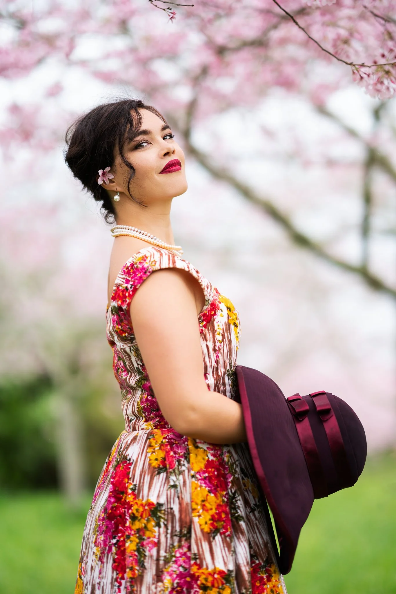 Anita in a floral dress holding a burgundy hat under cherry blossoms