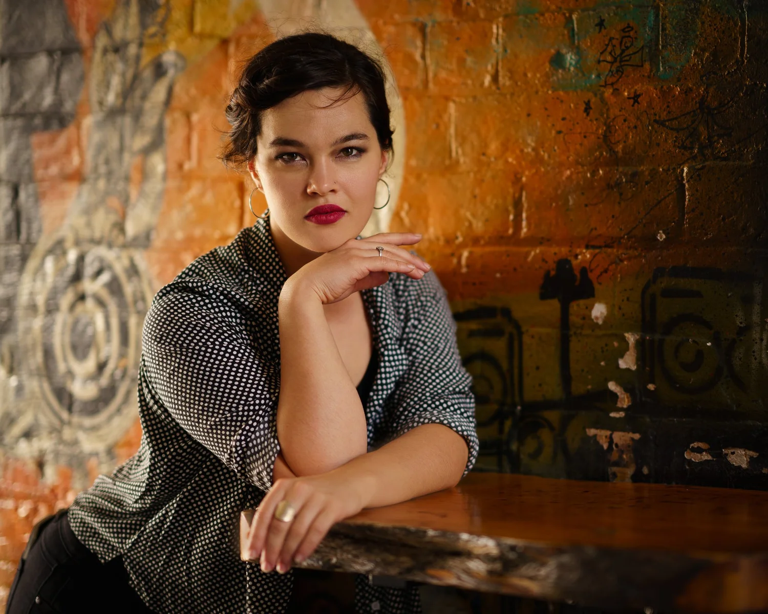 Anita leans on a wooden table against an orange brick wall