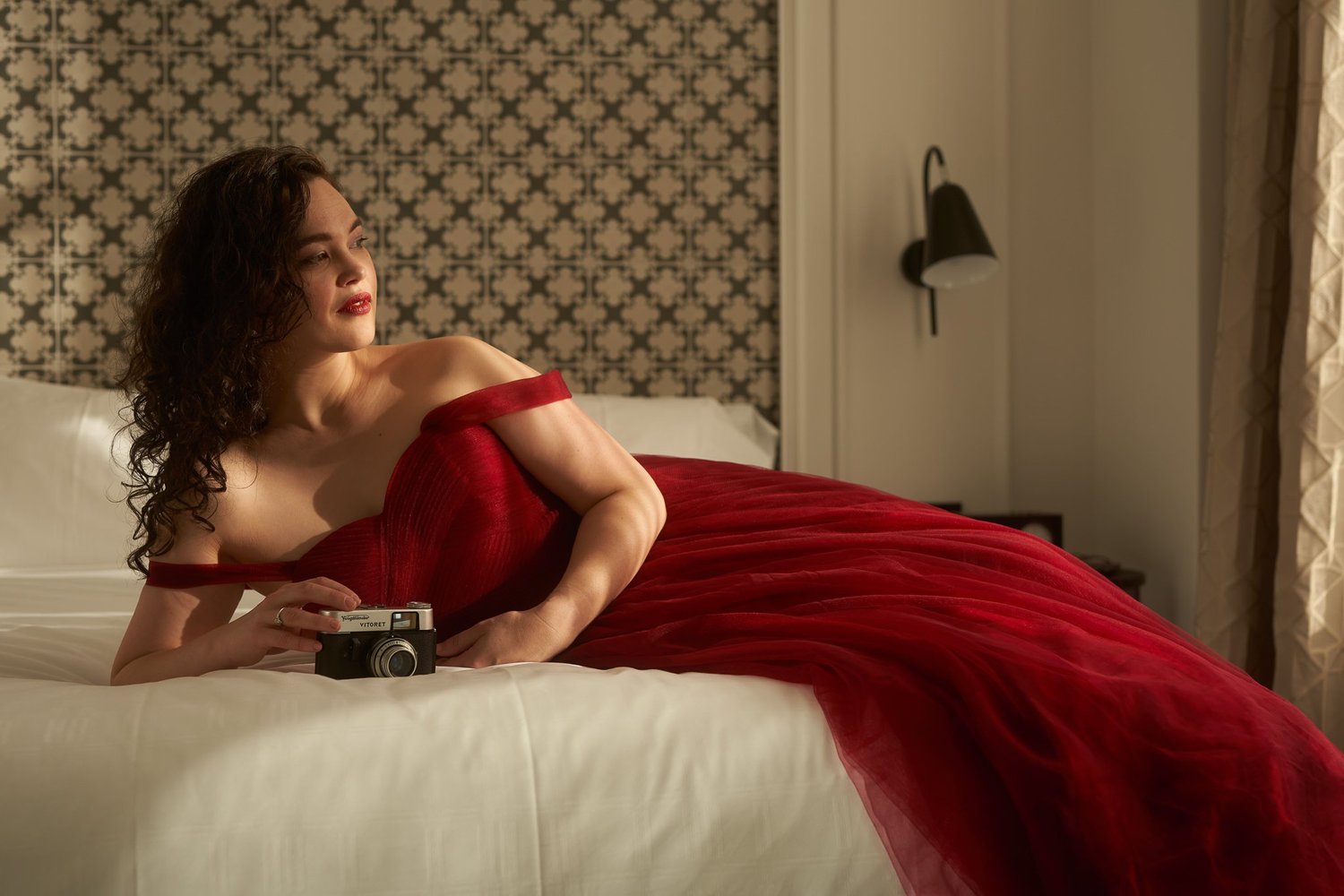 Anita in a red gown reclining on a hotel bed with a vintage camera