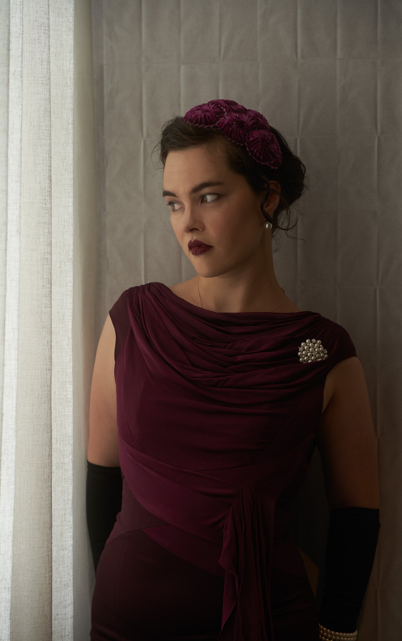 Anita in a burgundy dress and black gloves by a window