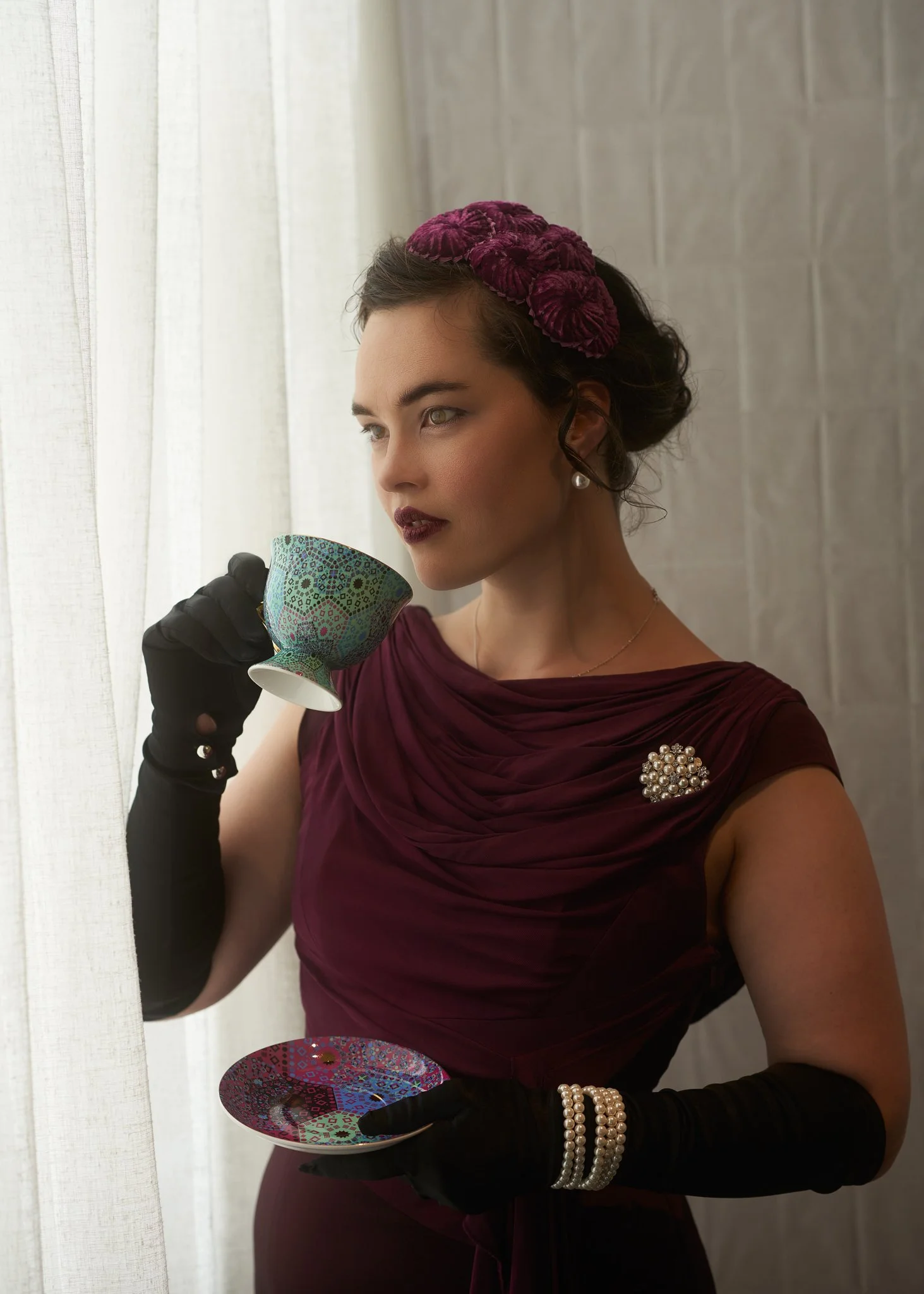 Anita in a burgundy dress and black gloves sipping from a teacup
