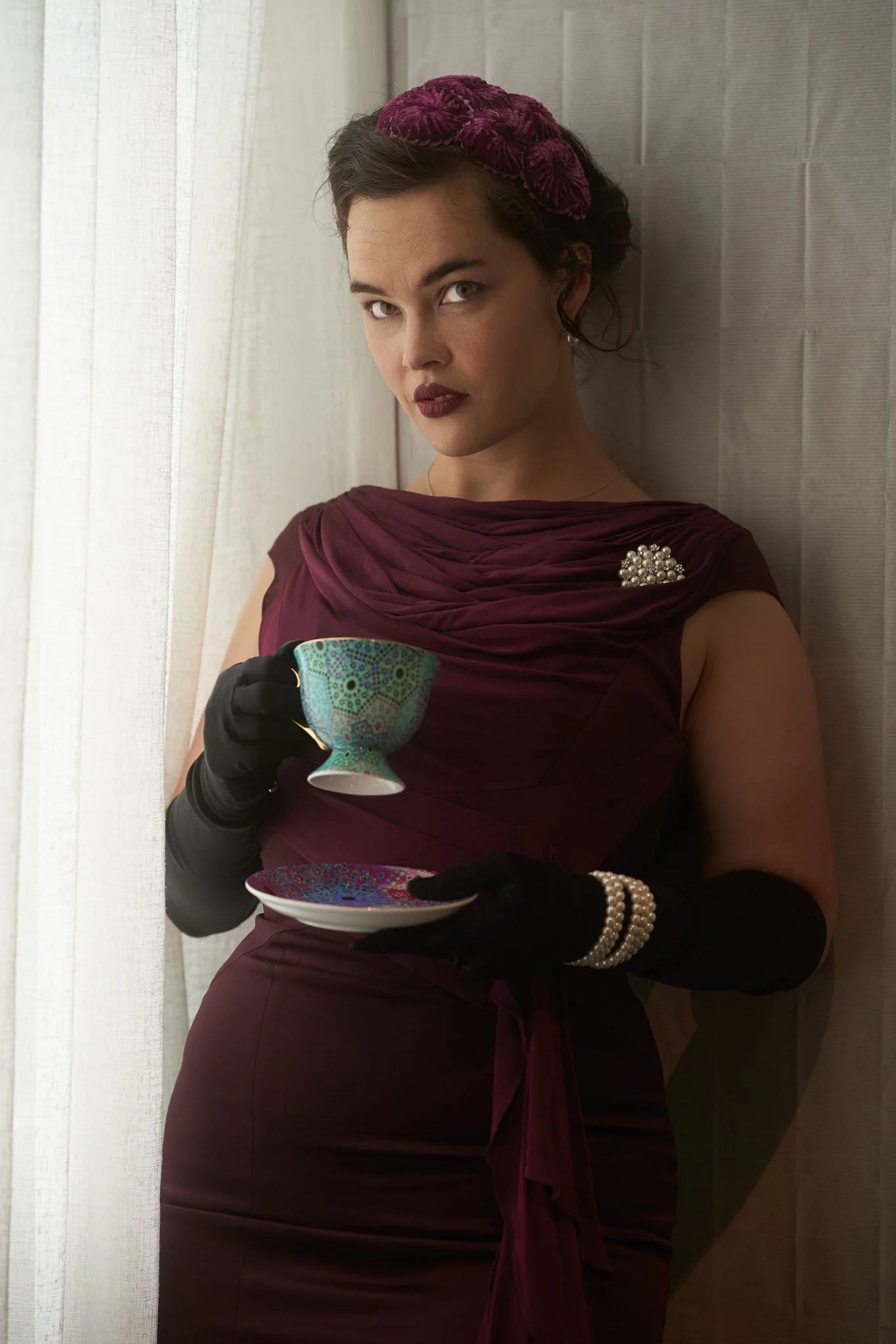 Anita holds a teacup and saucer in a burgundy dress