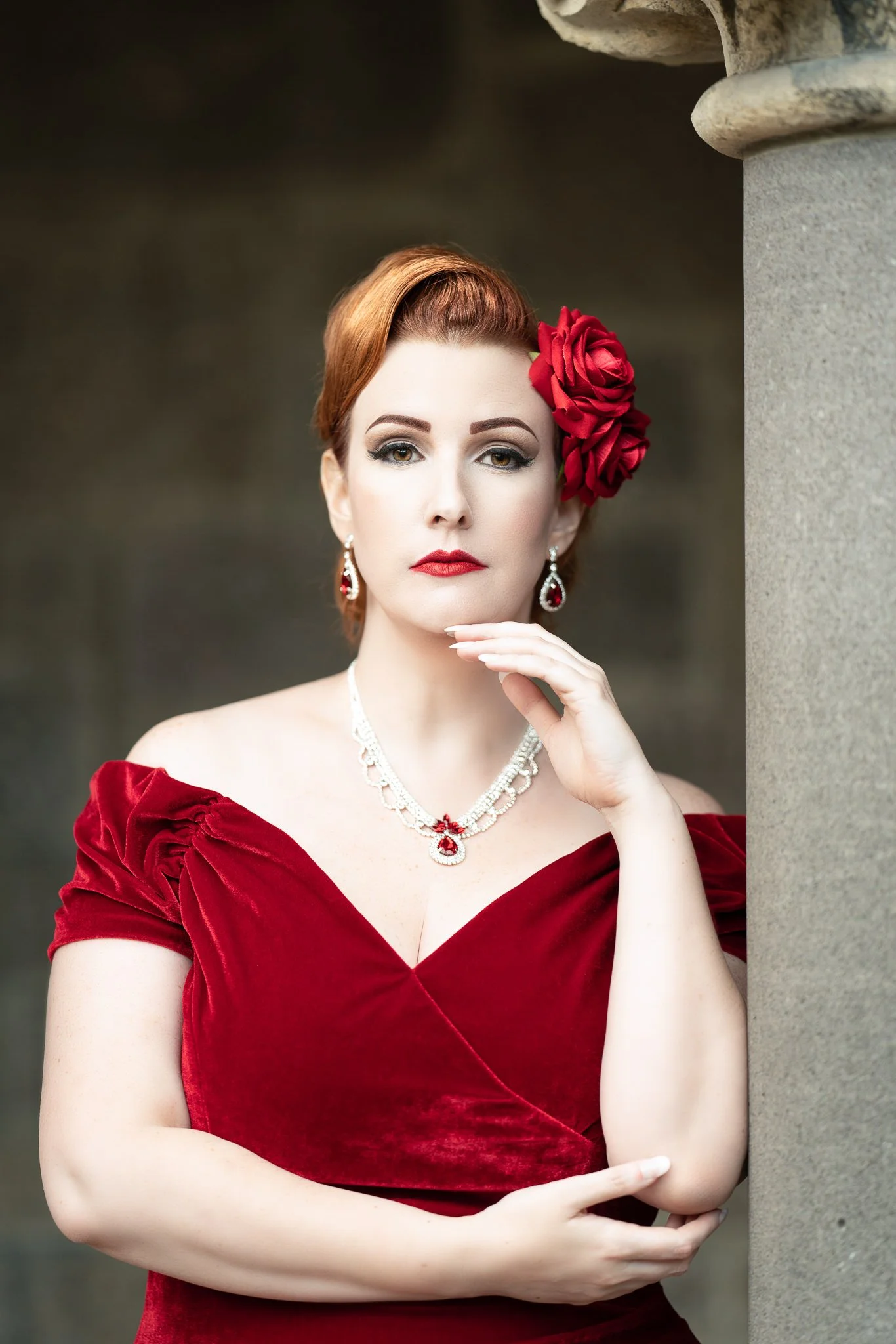 Jasmine in a red velvet off-the-shoulder dress beside a stone column