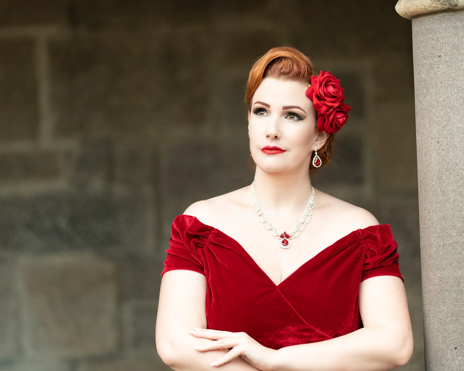 Jasmine in red velvet with arms folded against a stone wall