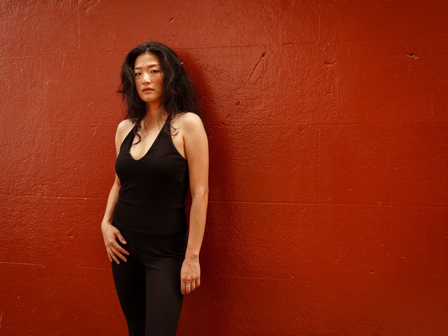 Anna stands against a red wall in an all-black outfit