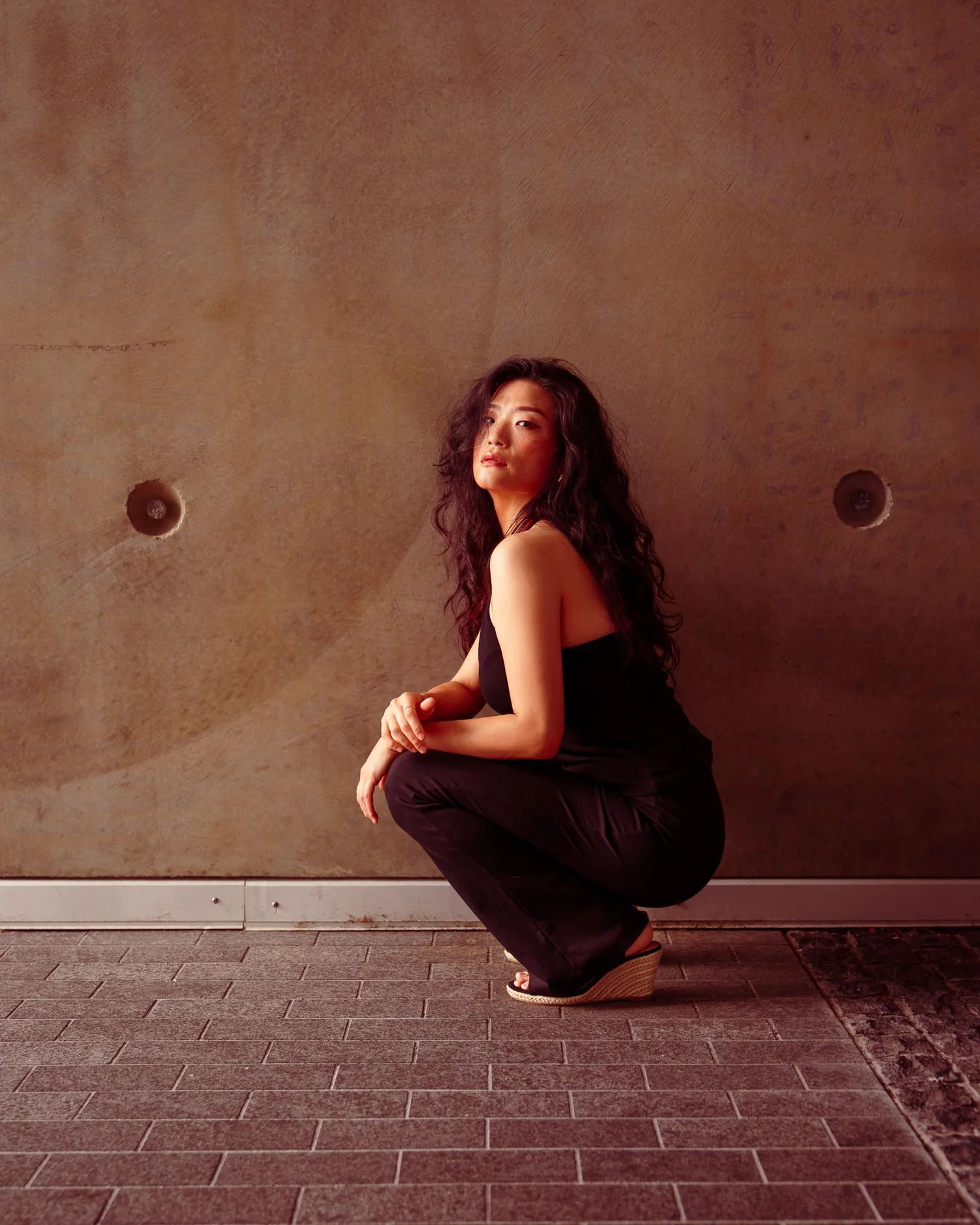 Anna crouches against a concrete wall looking away