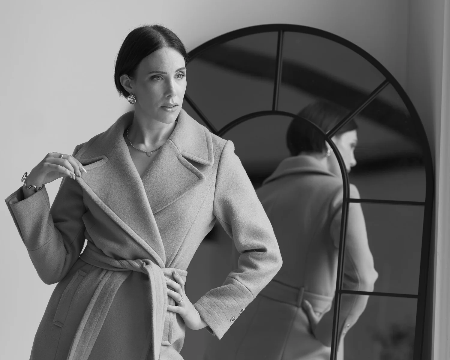 Black and white photo of Nat in a wrap coat beside an arched mirror