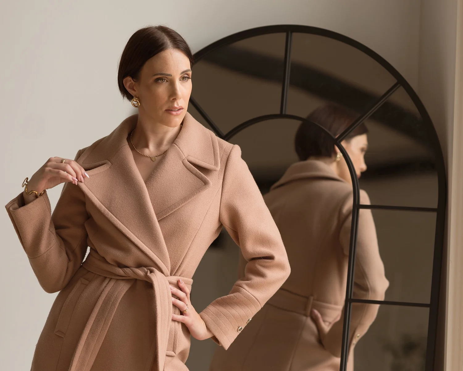 Nat Hugill in a camel coat in front of an arched mirror