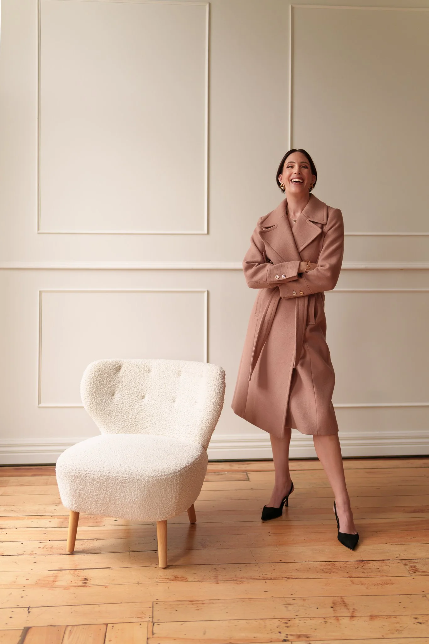 Nat laughs in a blush coat beside a white armchair