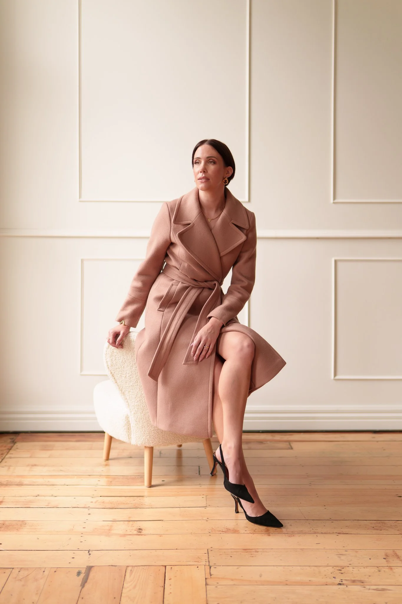 Nat sits on a white chair in a wrap coat and heels