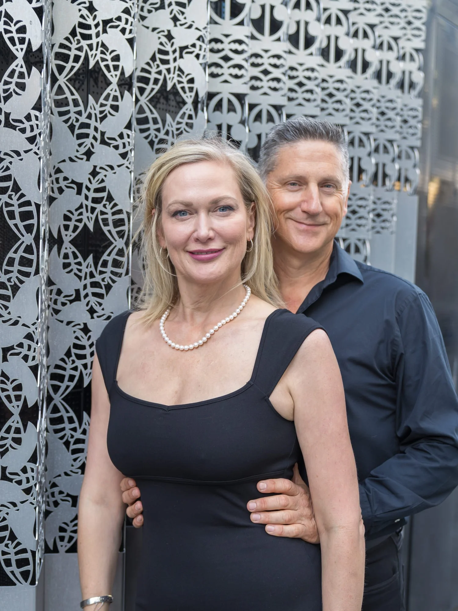 The couple smile in front of a patterned metal wall