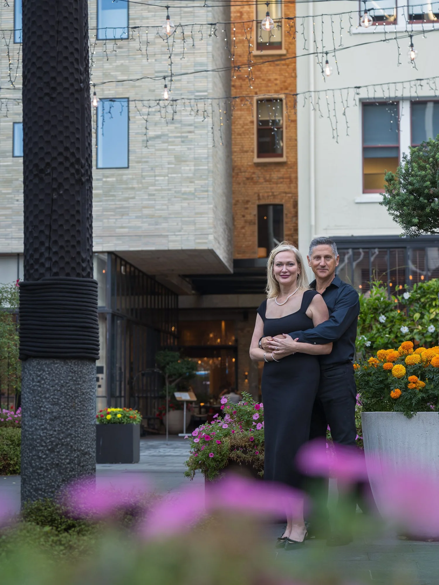 The couple stand framed by pink flowers in a laneway