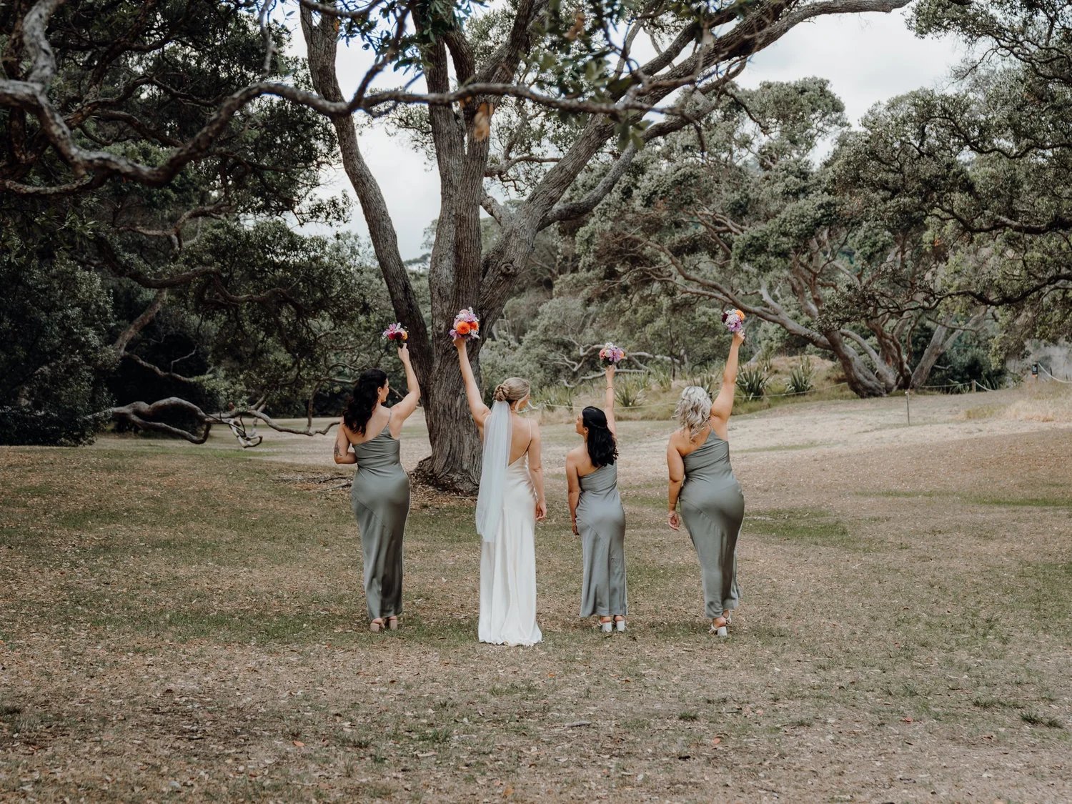 The bridesmaids raise their bouquets under a large tree
