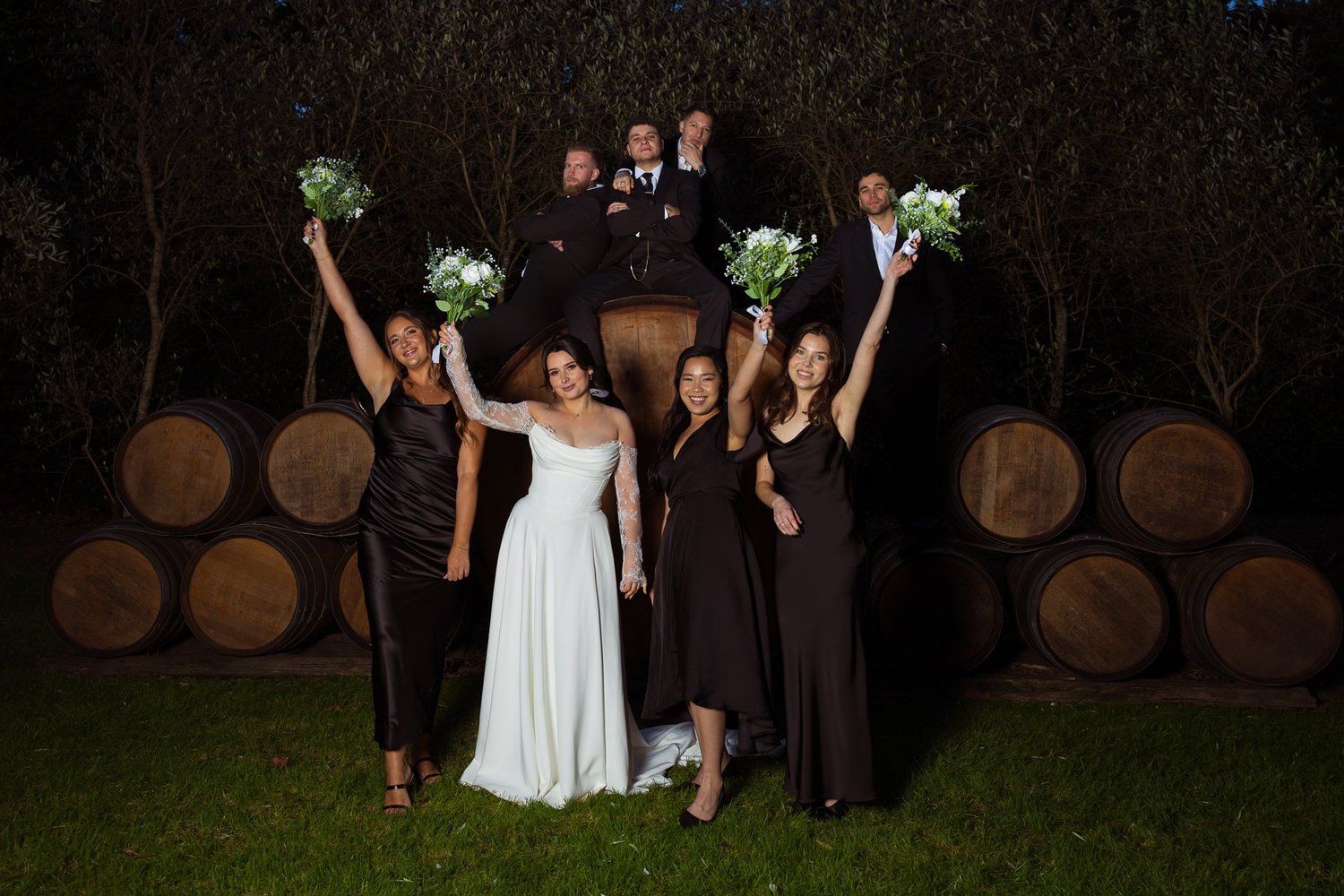 The bridesmaids raise their bouquets among the barrels at night