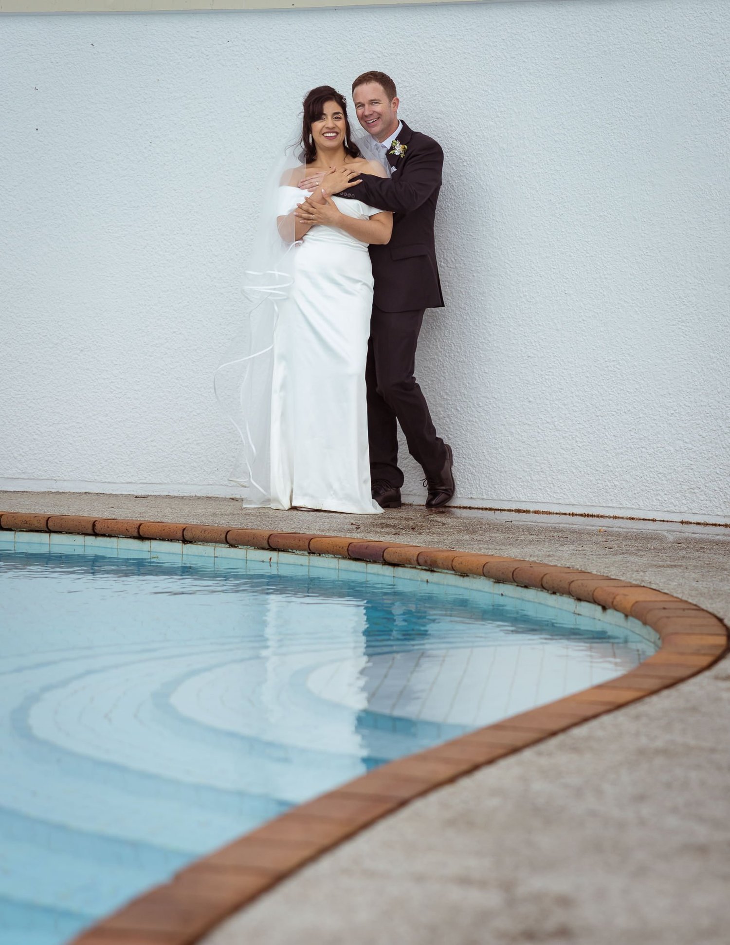 Feby and Steven lean against a white wall by the pool