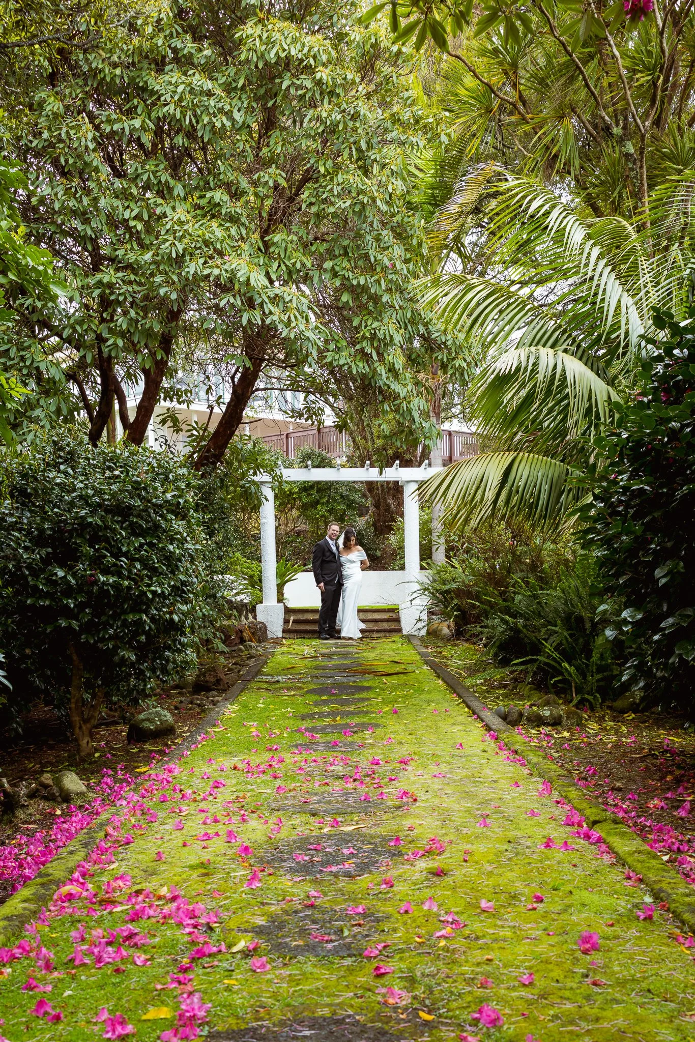 A moss-covered path scattered with pink petals leading to a white arbour