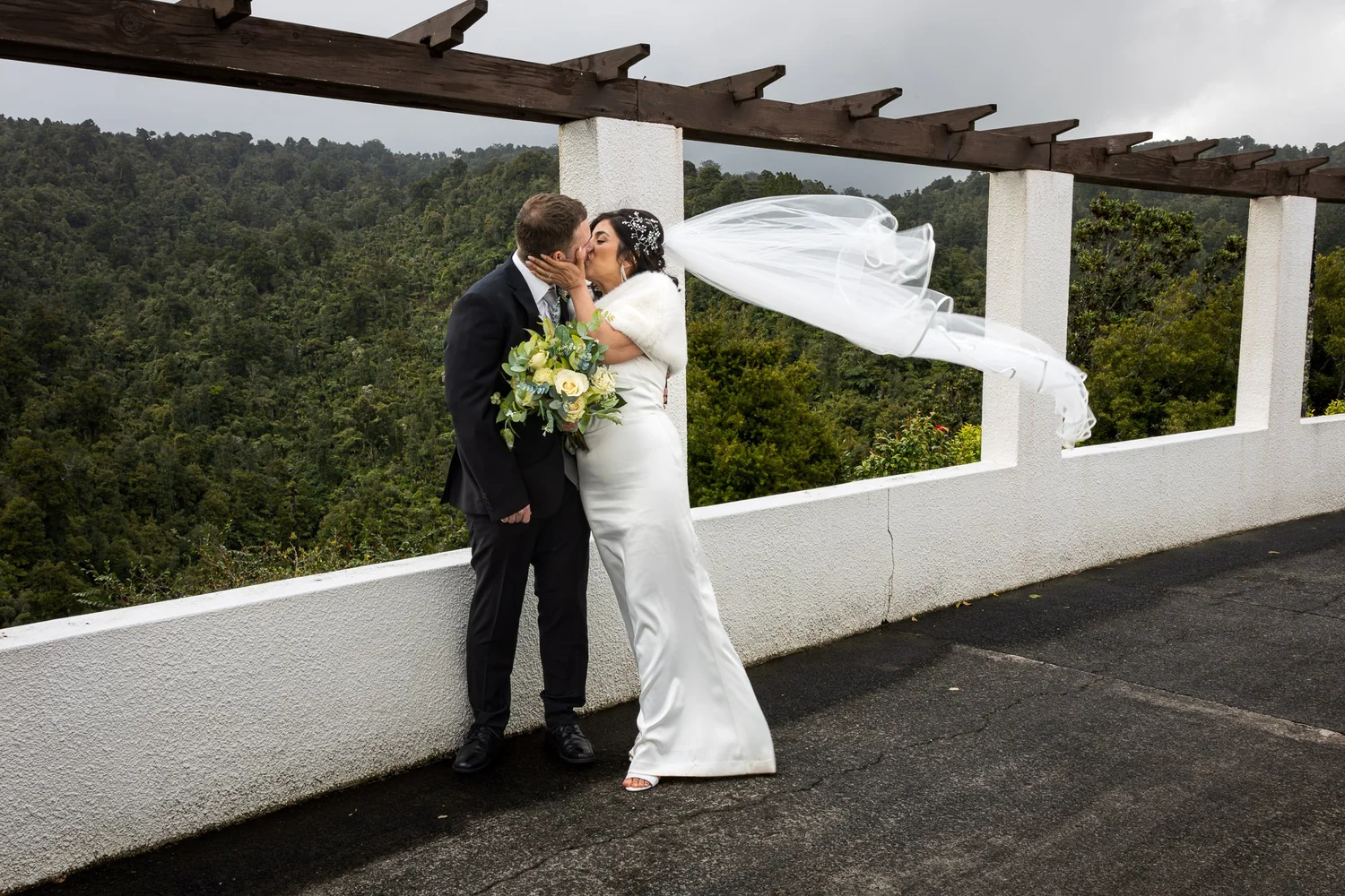 Feby's veil catches the wind as she kisses Steven above the bush at The Waitākere Resort and Spa